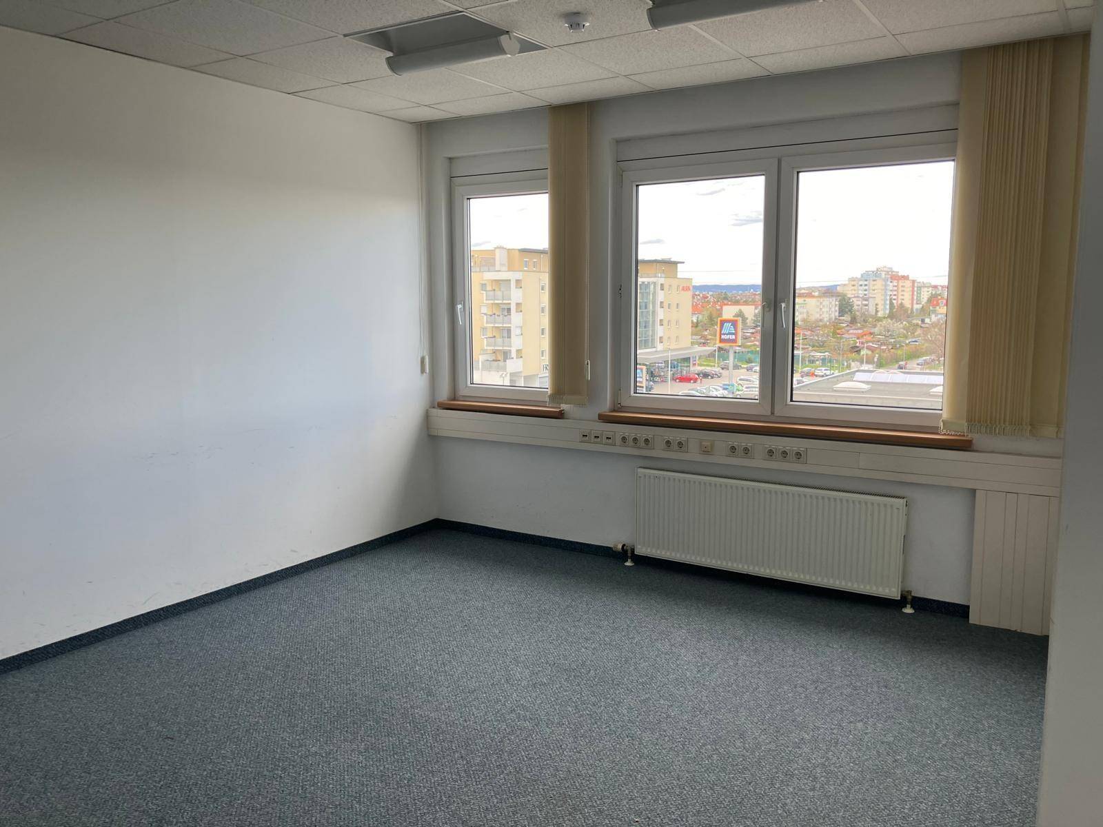 Leeres Büro mit Teppichboden, weißen Wänden und großen Fenstern mit Blick auf die Stadt.