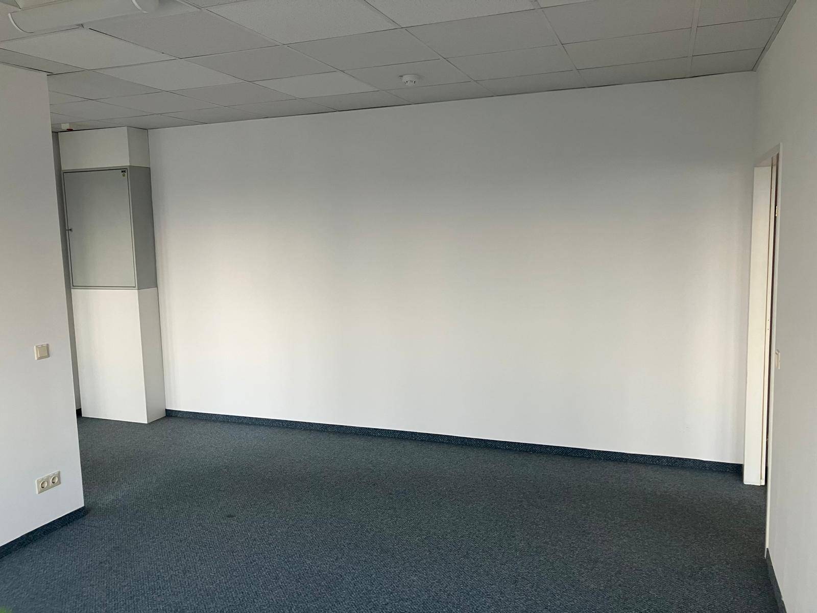 Heller Bürobereich mit Teppichboden und weißen Wänden, führt zu angrenzenden Räumen.