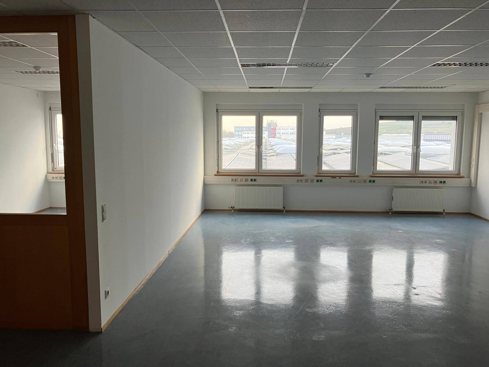 Leeres Büro mit drei Fenstern, die einen Blick auf Dächer mit Photovoltaikanlagen bieten.