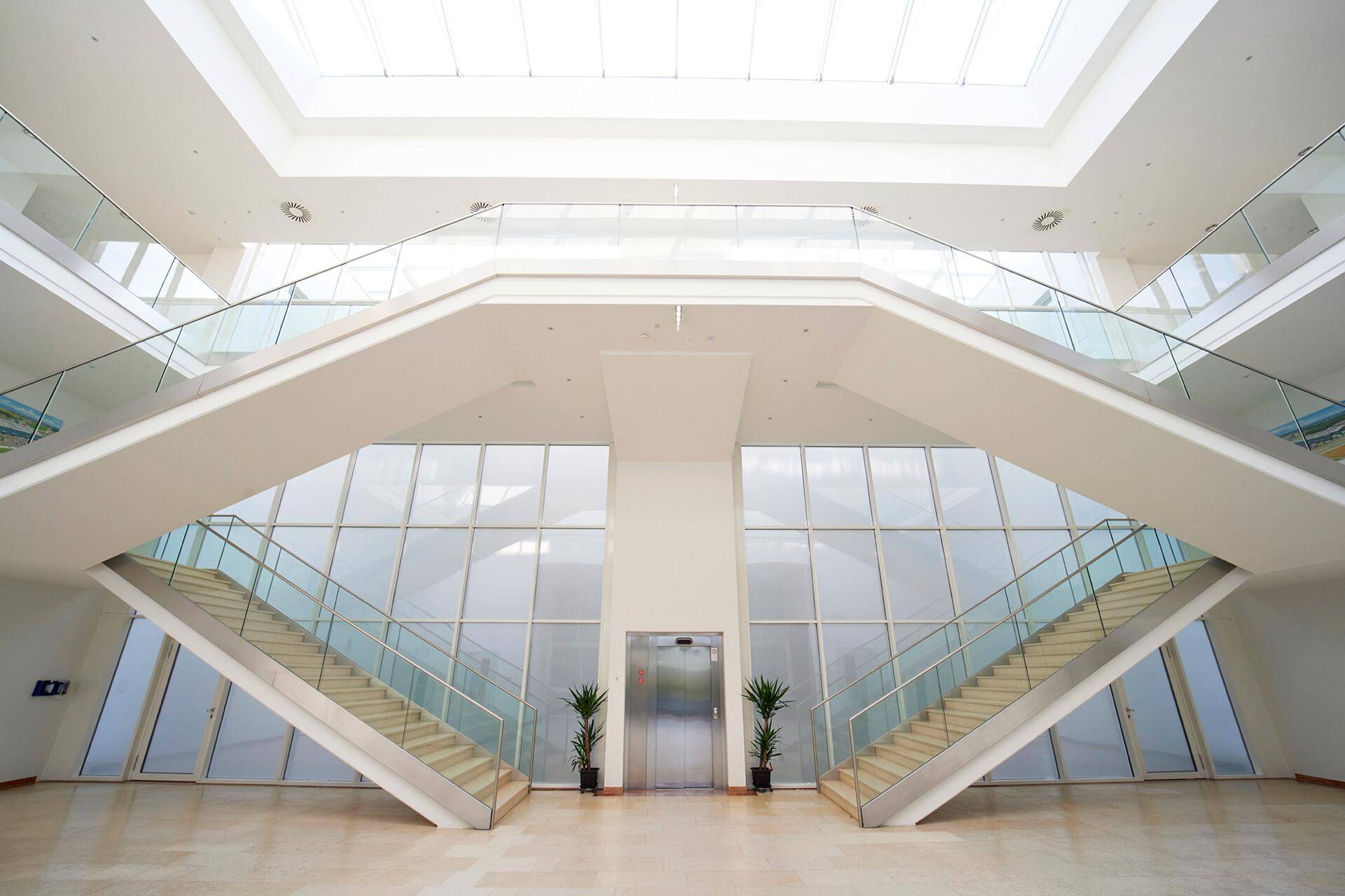 Helle, moderne Lobby mit beeindruckender symmetrischer Treppe, Glasgeländern und zentralem Aufzug unter einem Oberlicht.