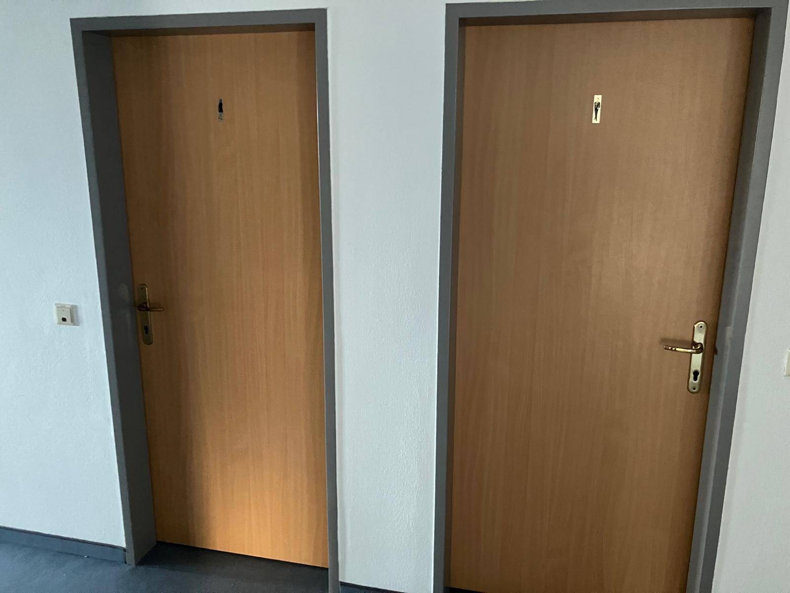 Zwei schlichte Holztüren in einem Flur, gekennzeichnet für Damen- und Herrenbereiche, mit grauen Türrahmen.