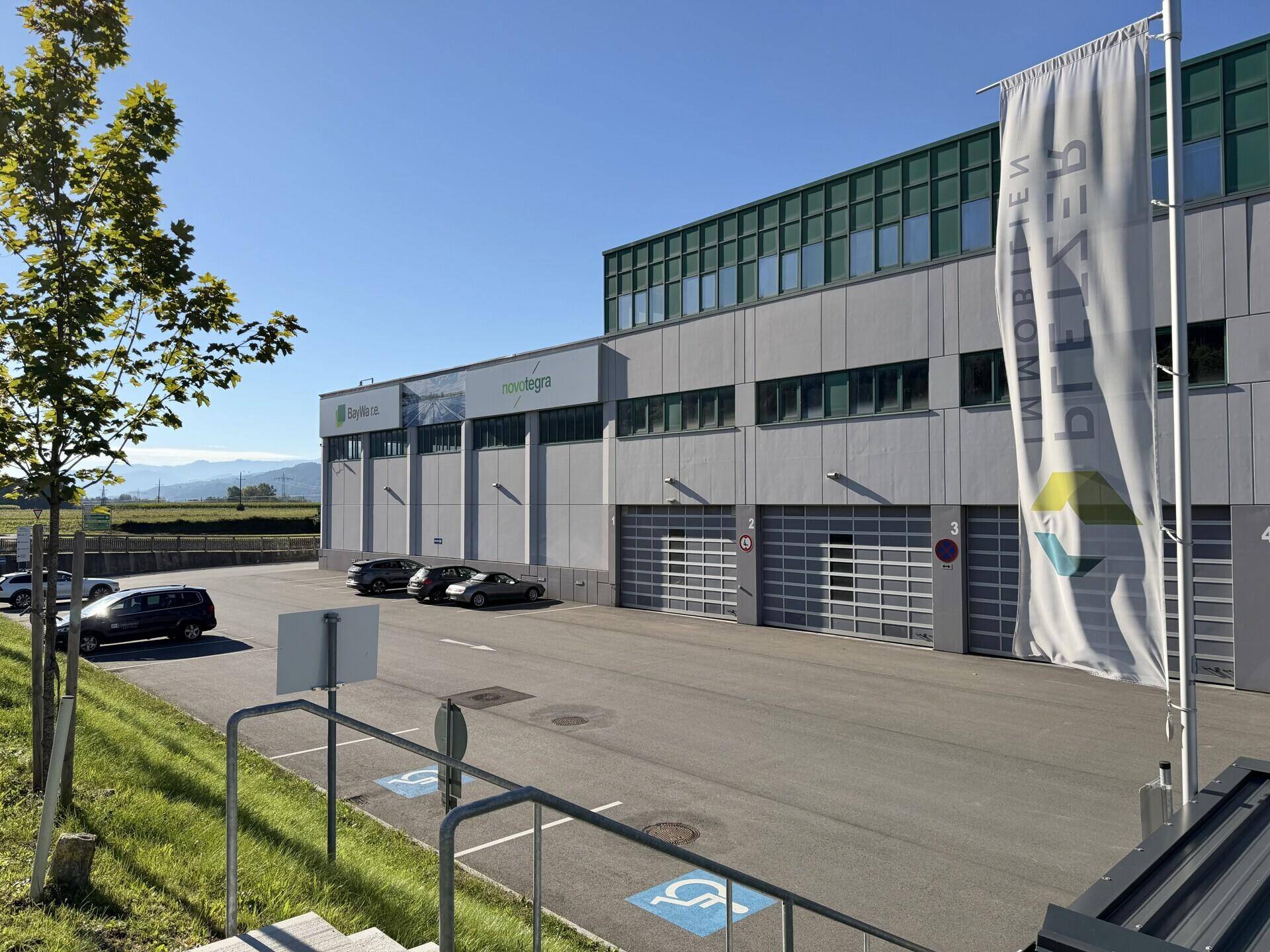 Modernes Gewerbegebäude mit Parkplätzen, Laderampen und einer Fahne vor dem Eingangsbereich.
