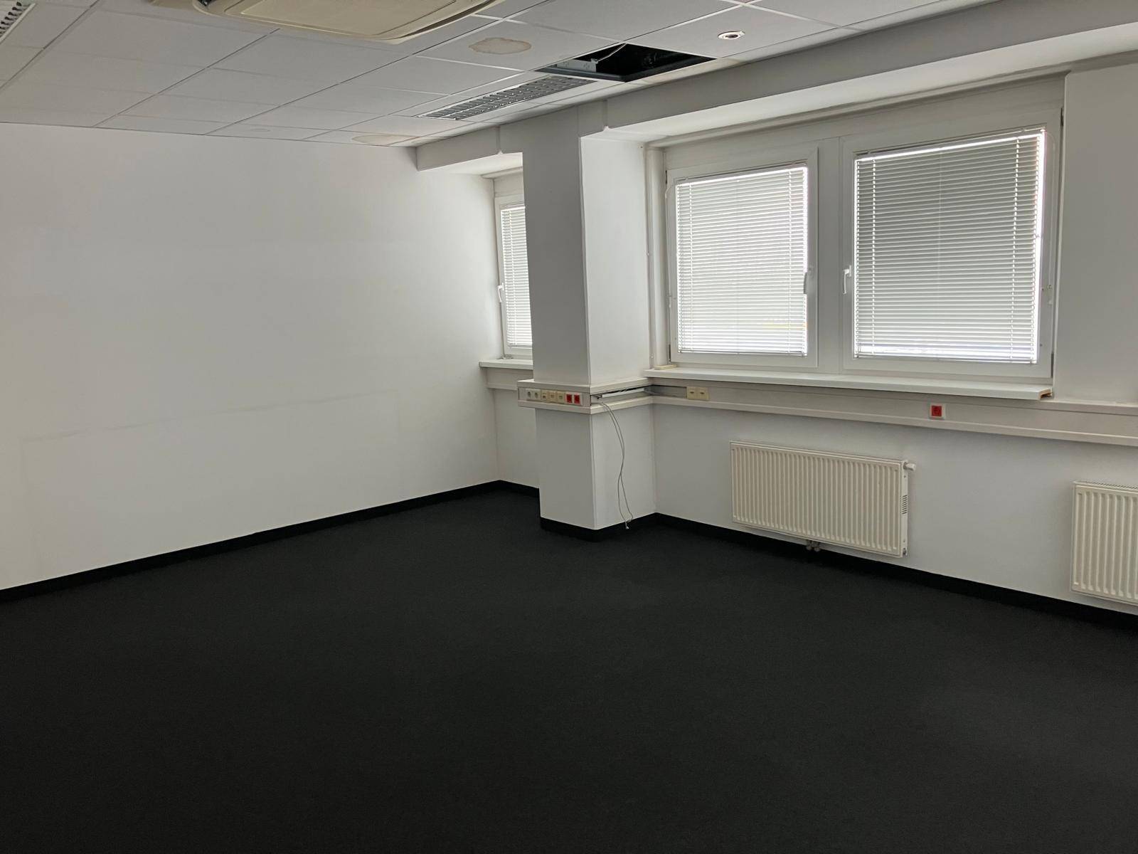 Geräumiges Büro mit großen Fenstern, die viel Tageslicht hereinlassen, und dunklem Teppichboden.