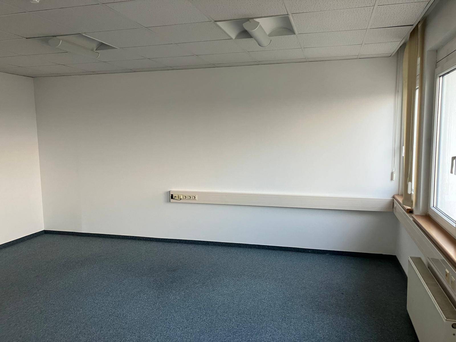 Büroraum mit Teppichboden, weißen Wänden, Steckdosenleiste und Fenster.