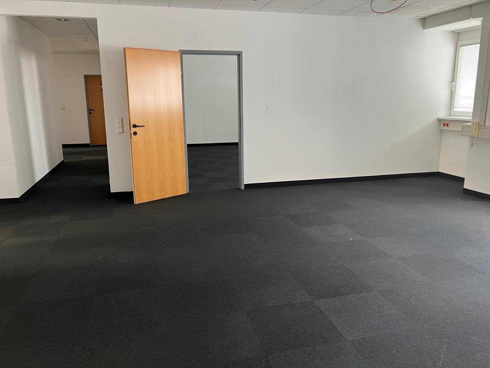 Geräumiges, leeres Büro mit dunklem Teppichboden und mehreren Türen, bereit zur individuellen Gestaltung.
