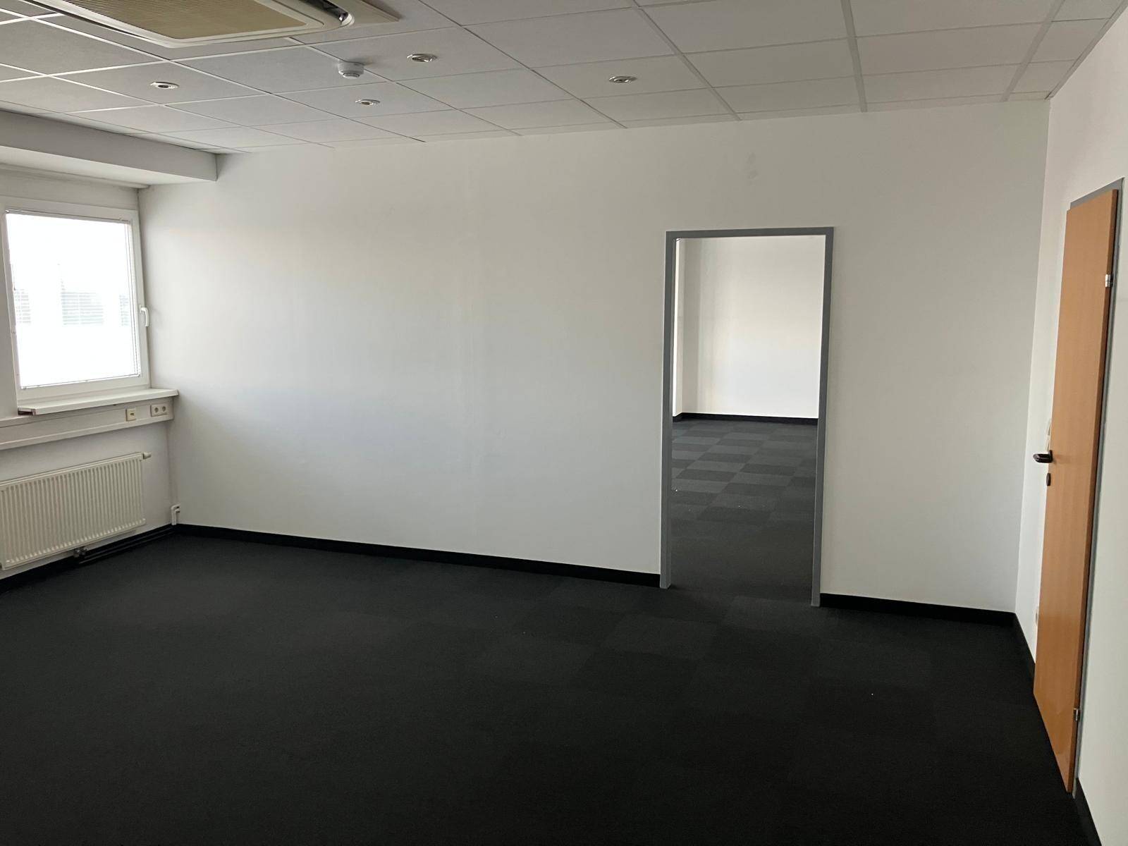 Leeres Büro mit dunklem Teppichboden, einem Fenster und einer Holztür, bereit zur individuellen Gestaltung.