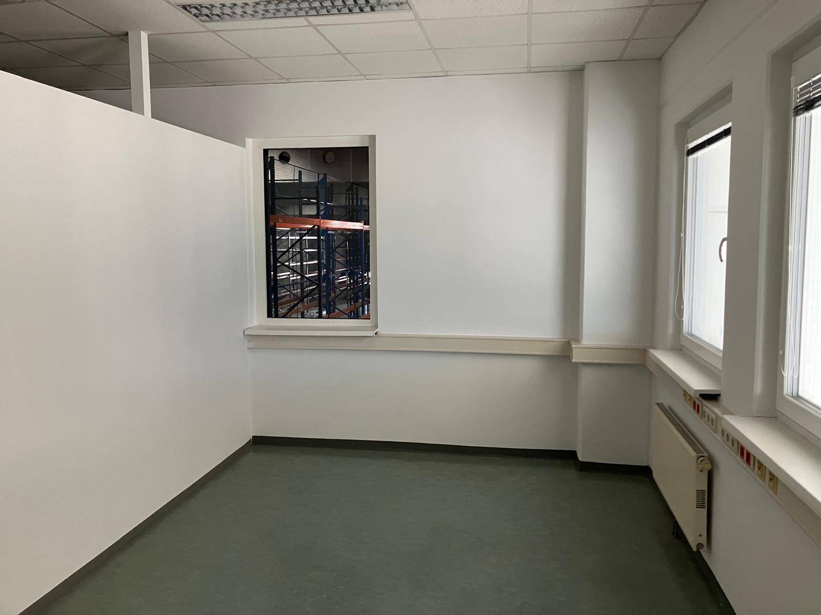 Büro mit weiß gestrichenen Wänden, einem Fenster zur Lagerhalle und einem dunklen Vinylboden, sauber und funktional.
