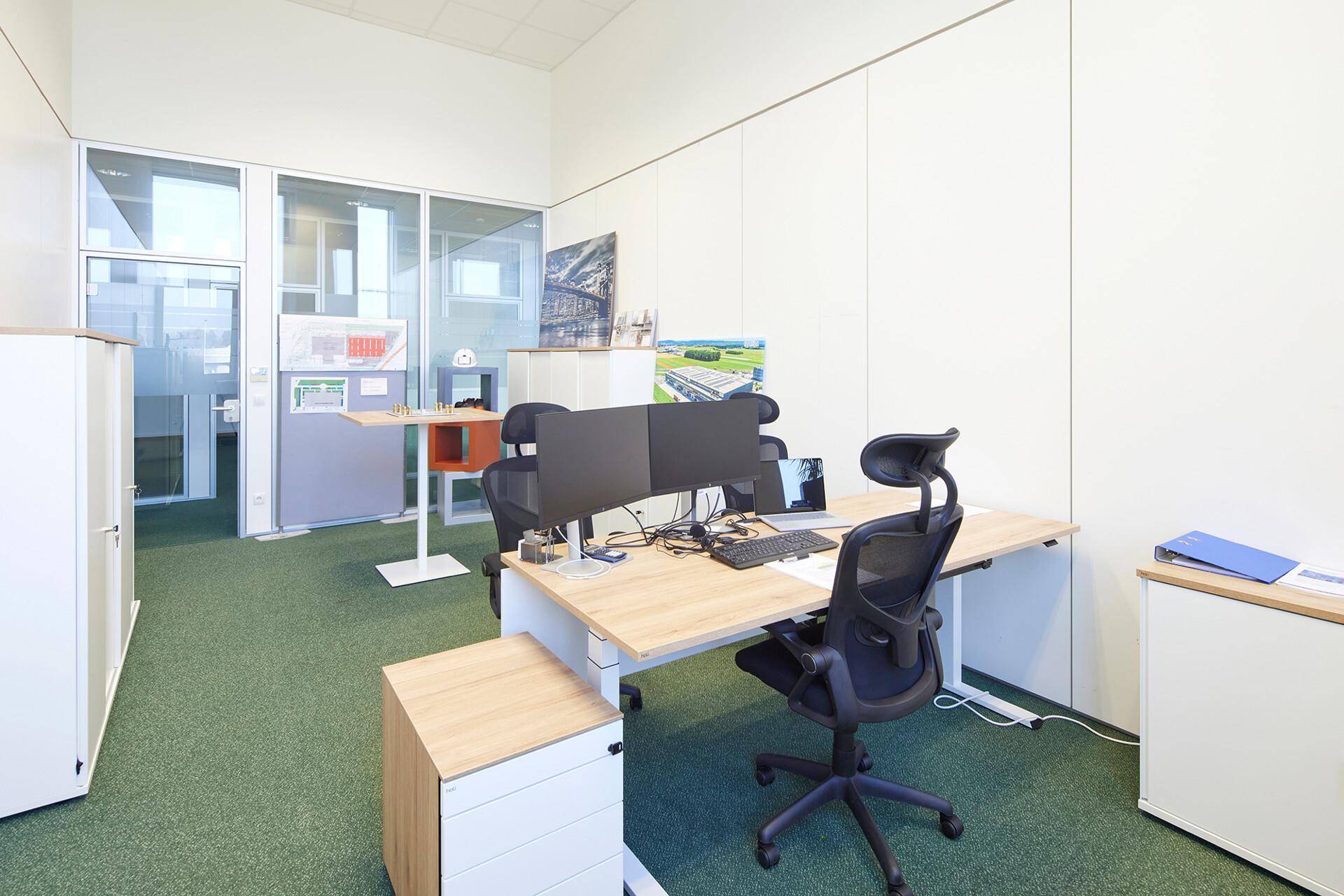 Modernes Büro mit grünem Teppichboden, höhenverstellbarem Schreibtisch und Glaswänden zu angrenzenden Räumen.