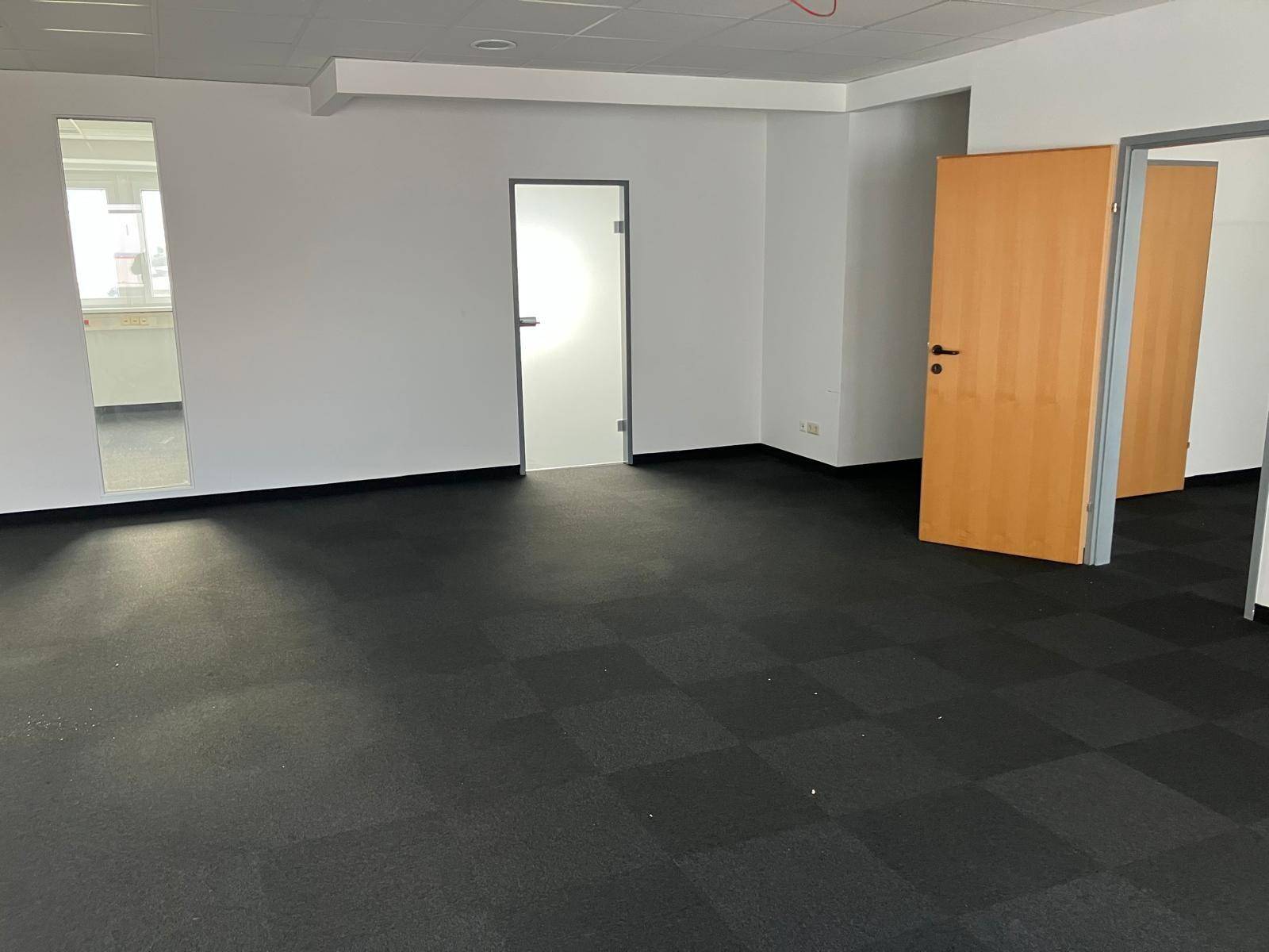 Großes, leeres Büro mit dunklem Teppichboden und verschiedenen Türen, darunter eine moderne Glastür.