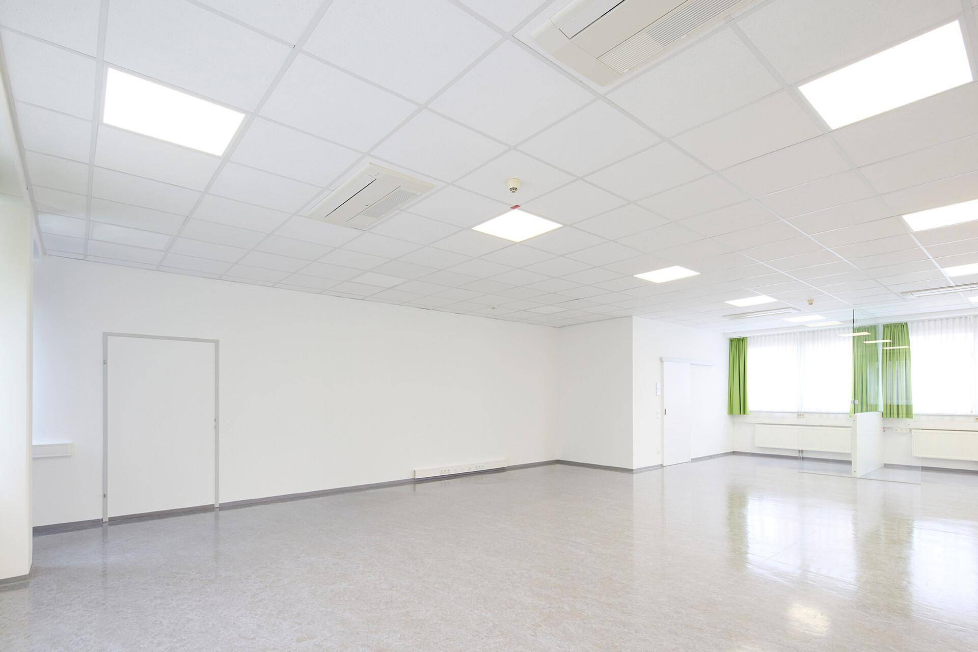 Großer, leerer Raum mit hellem Vinylboden, weißen Wänden und Klimaanlagen an der Decke.