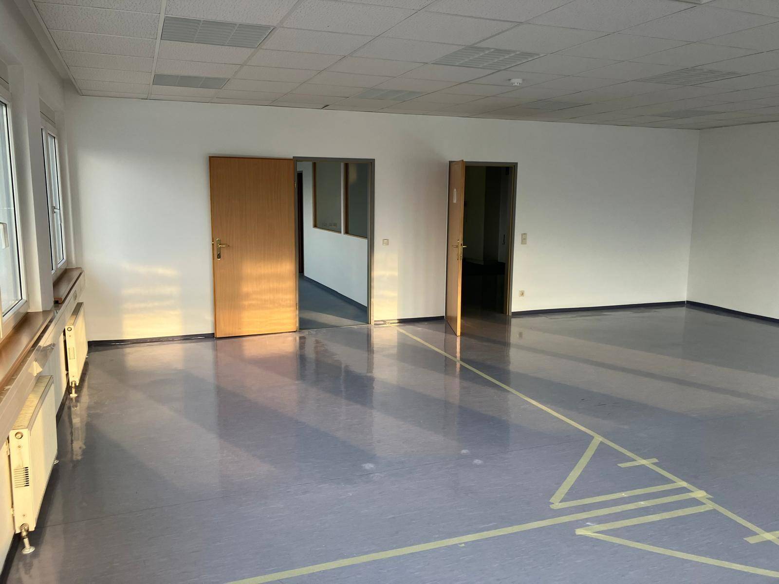 Geräumiger, leerer Büroraum mit großen Fenstern, Heizkörpern und hellem Boden, durchflutet von natürlichem Sonnenlicht.