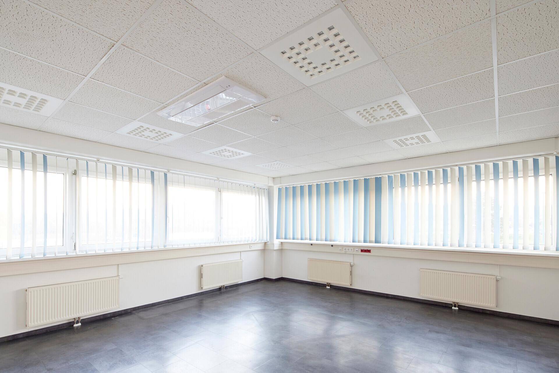 Leeres Büro mit abgehängter Decke, integrierter Beleuchtung und großen Fenstern mit vertikalen Jalousien.
