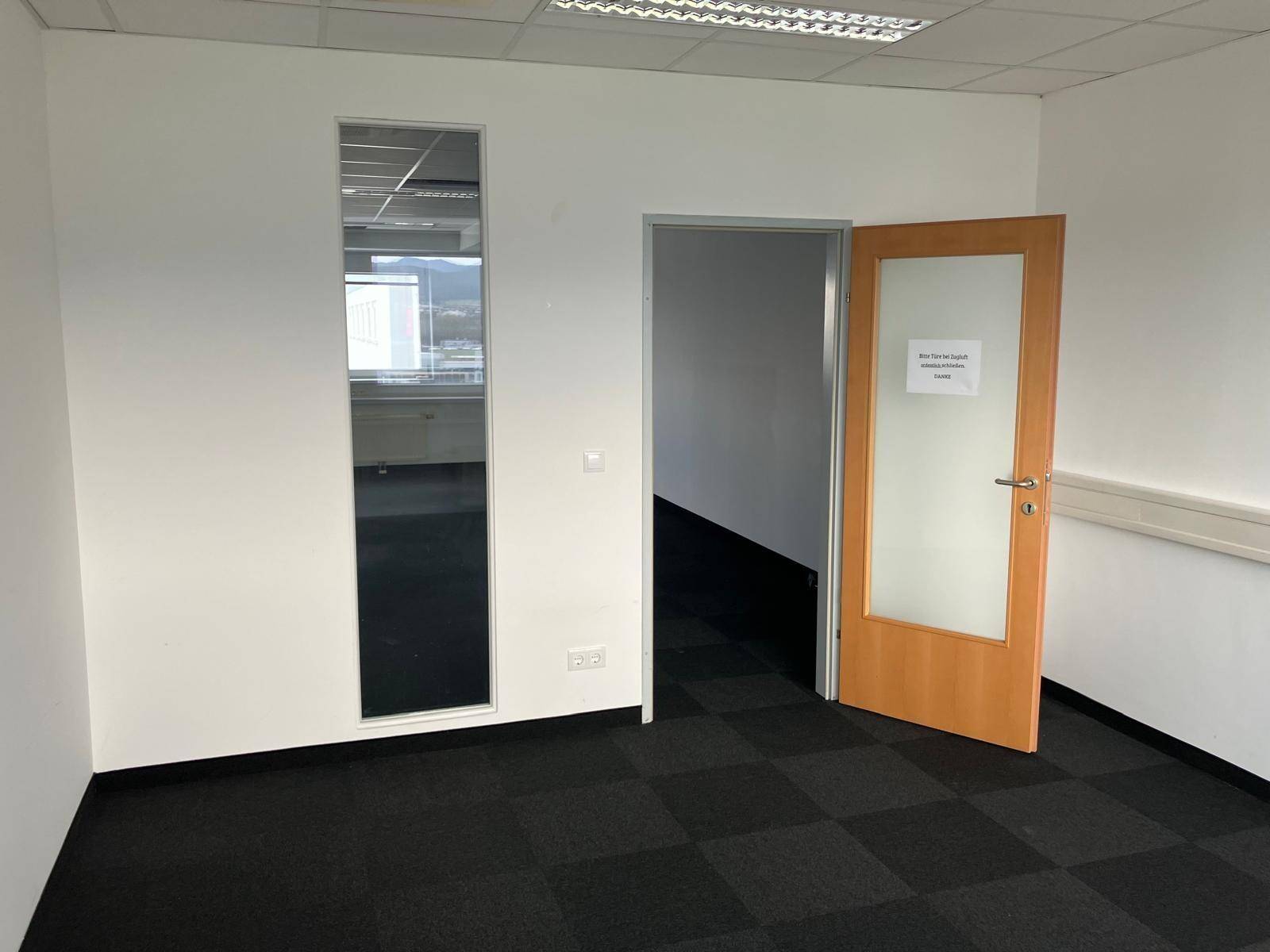 Geräumiges, leeres Büro mit dunklem Teppichboden und einer Holztür mit Milchglas, bereit zur Nutzung.