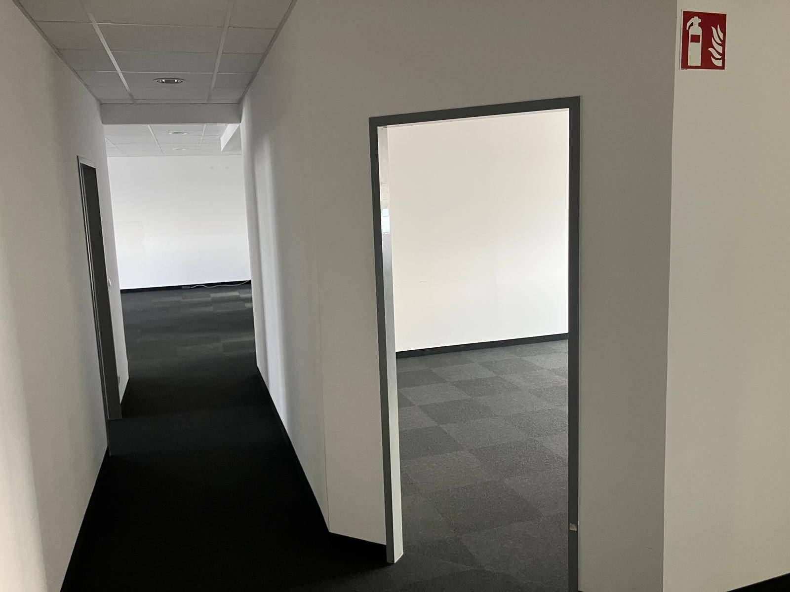 Langer, leerer Flur mit dunklem Teppichboden und mehreren Türen, der zu verschiedenen Büroräumen führt.