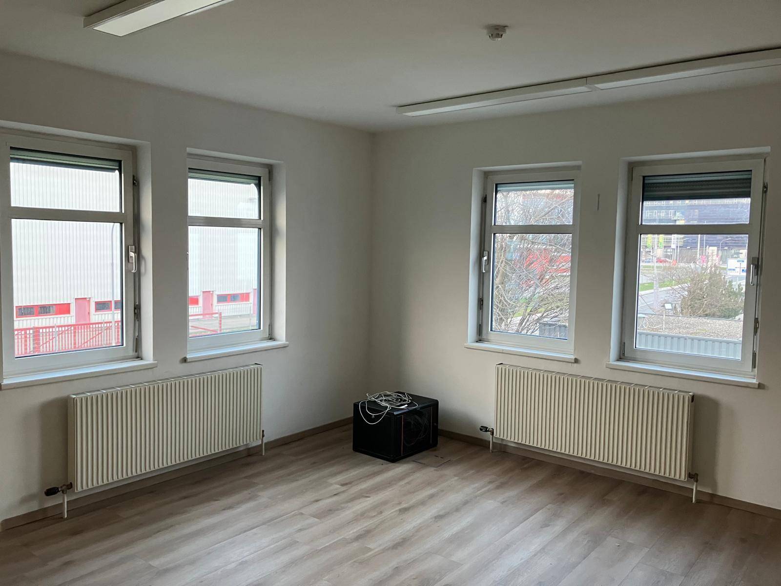 Geräumiger Büroraum mit vier Fenstern, Laminatboden und Heizkörpern, bietet viel Tageslicht.