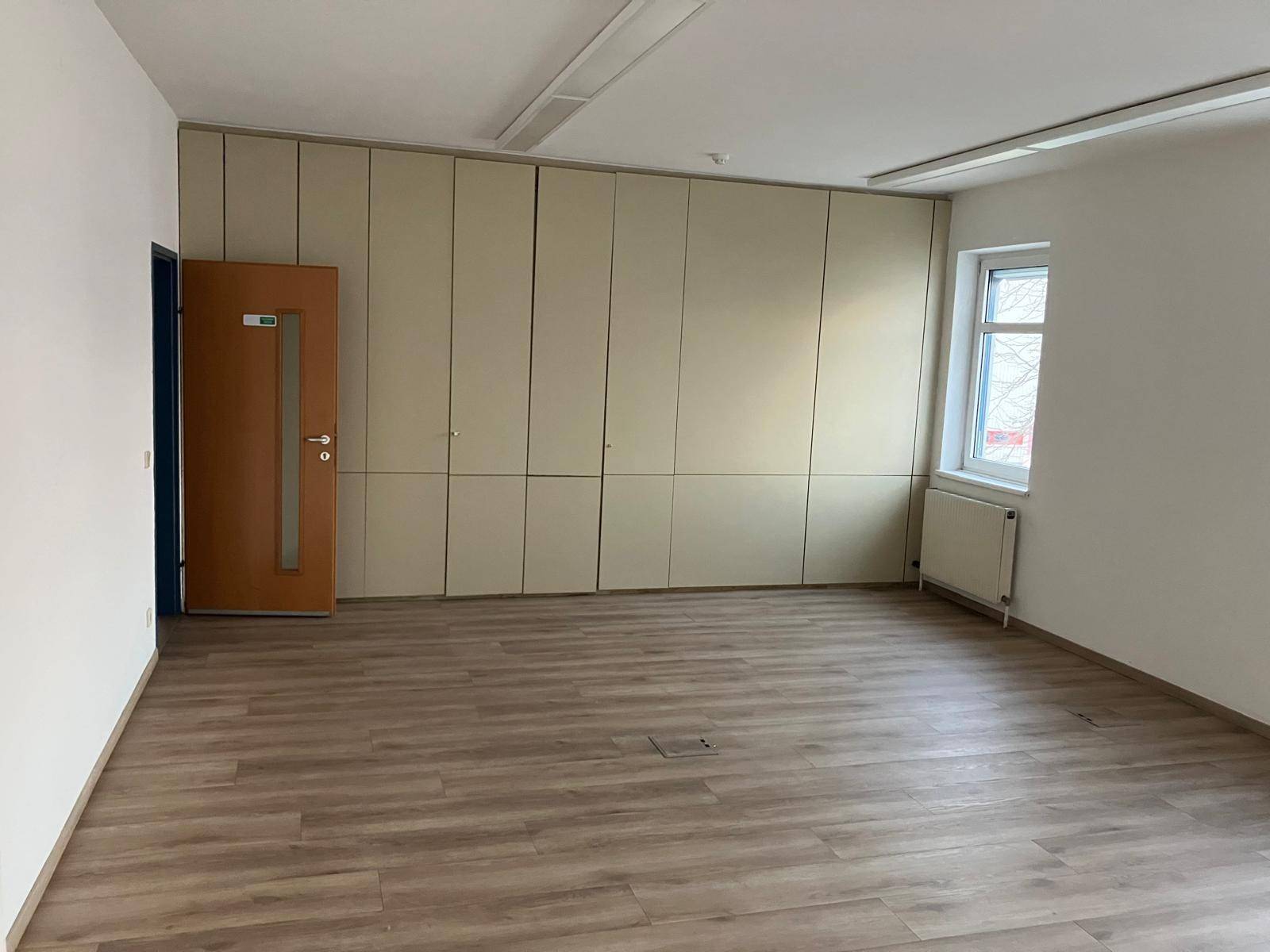 Heller Büroraum mit integrierten Schränken, Laminatboden und einem Fenster für Tageslicht.