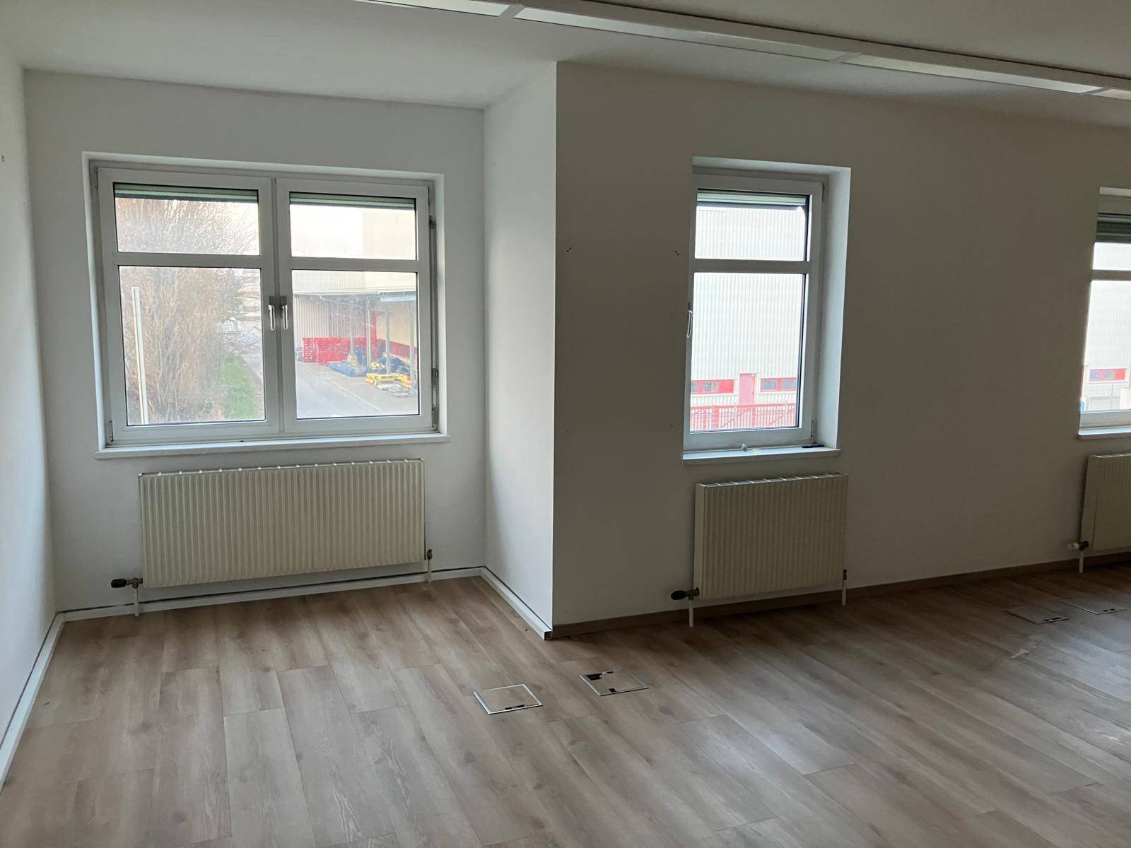 Geräumiger Büroraum mit zwei großen Fenstern, Laminatboden und Heizkörpern, bietet eine helle Atmosphäre.