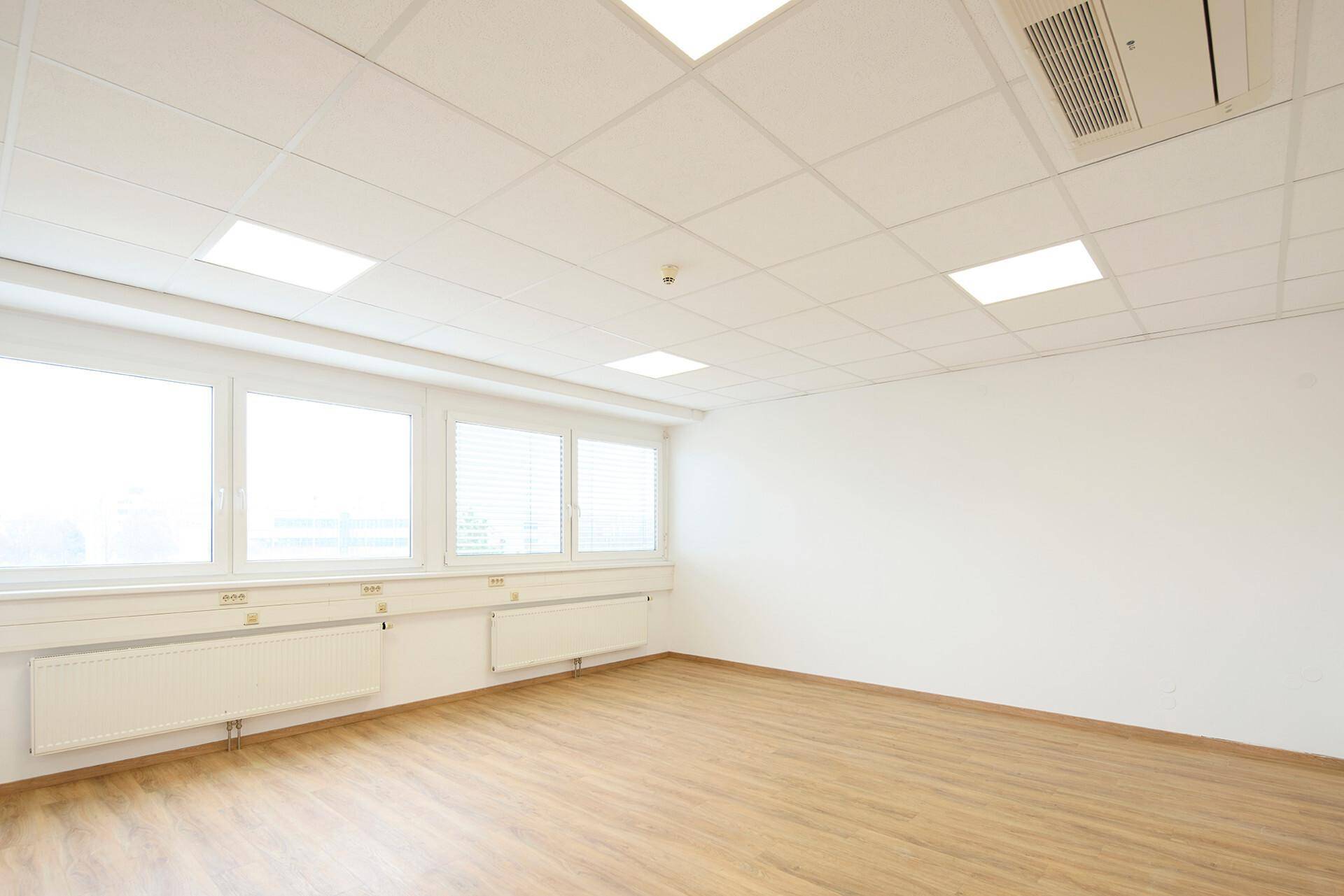 Leerer Büroraum mit Laminatboden, großen Fenstern, Heizkörpern und einer Klimaanlage an der Decke.