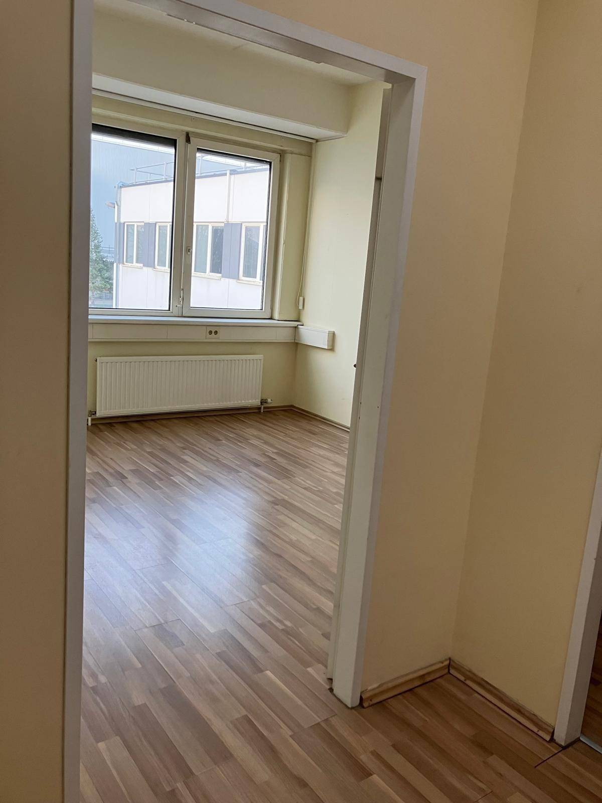 Leeres Büro mit großen Fenstern, Laminatboden und einem Heizkörper unter der Fensterbank, bietet grünen Ausblick.