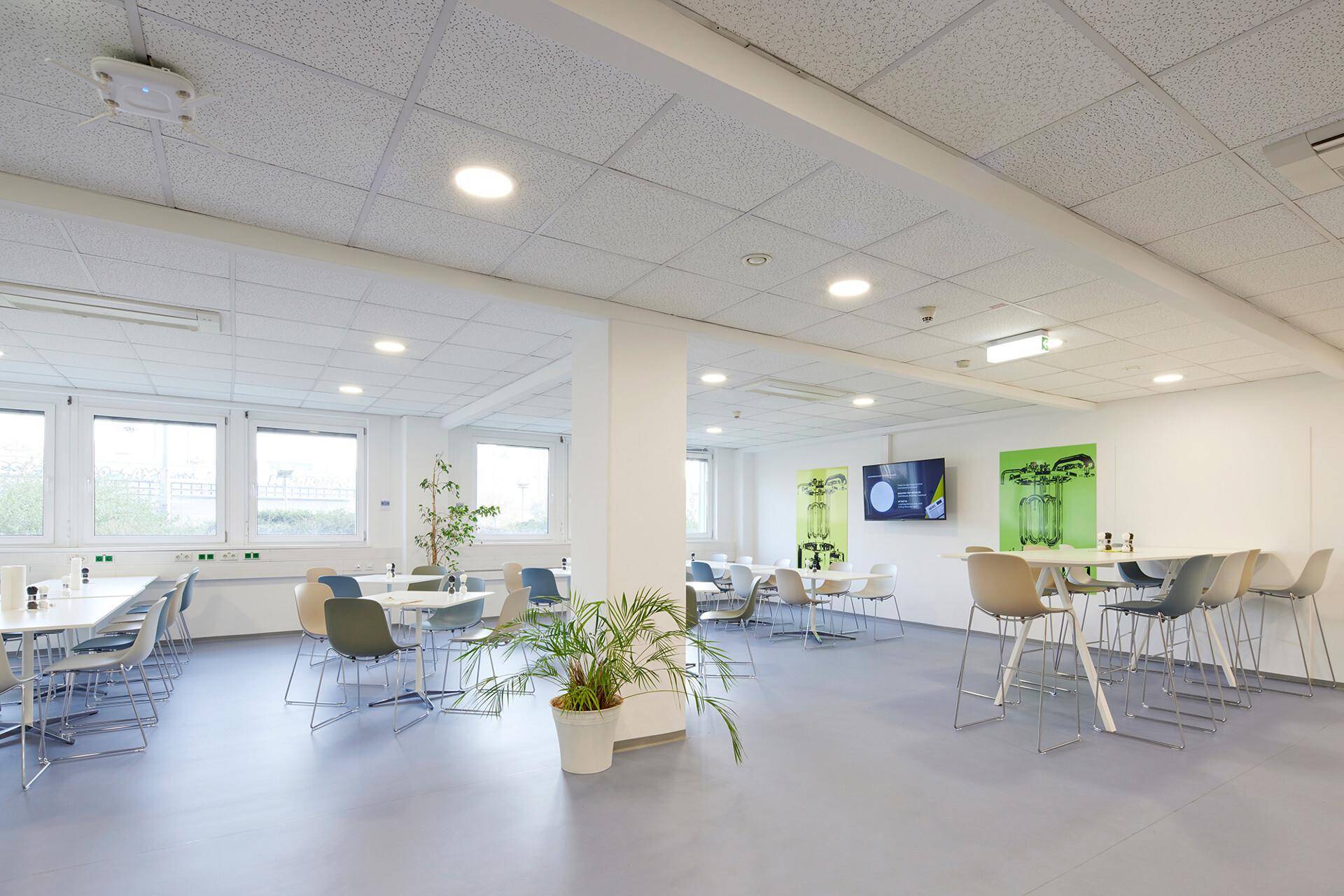 Geräumige Kantine mit mehreren Tischen und Stühlen, Pflanzen und großen Fenstern, die viel Licht hereinlassen.
