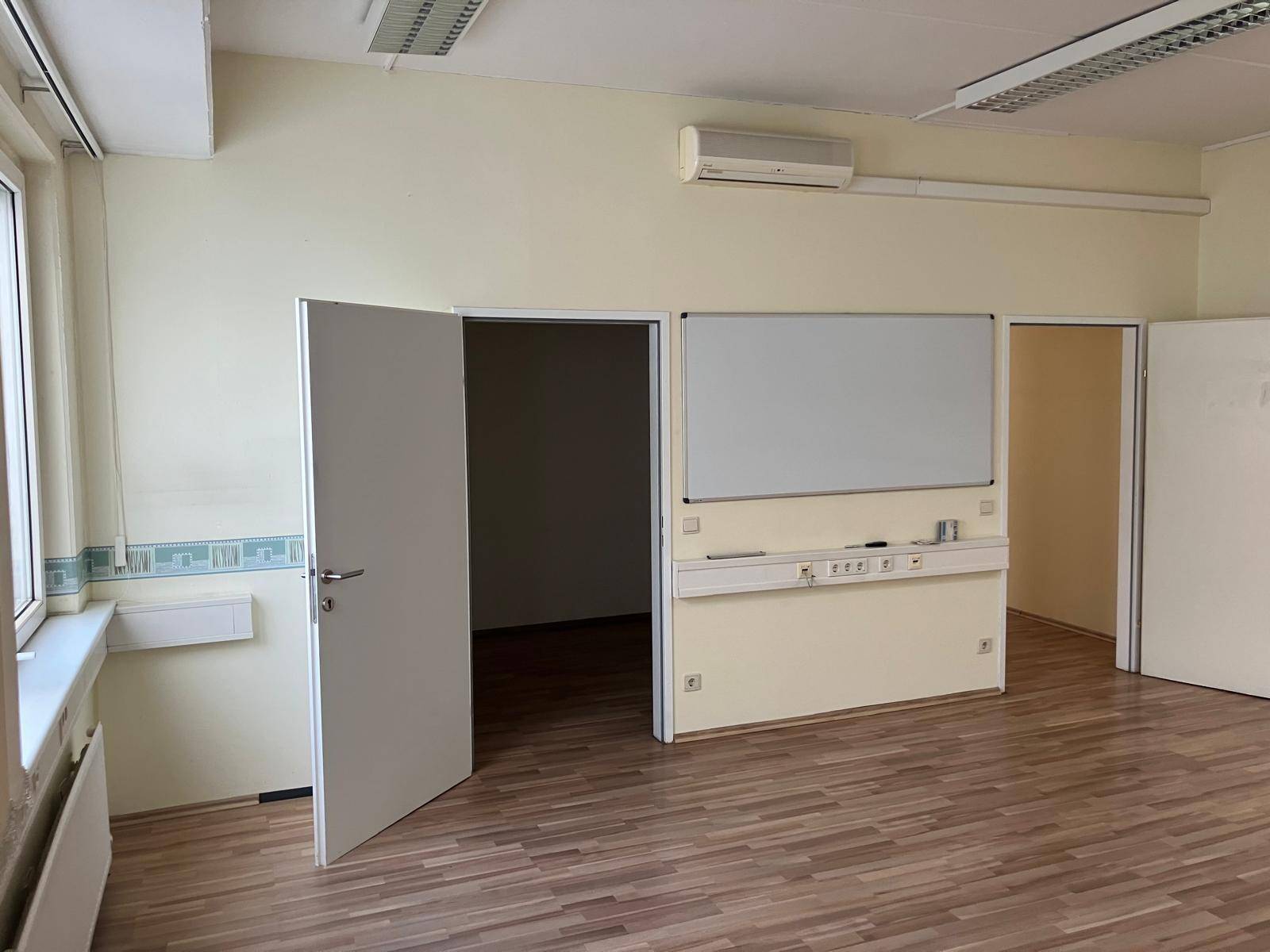 Geräumiges Büro mit Laminatboden, Klimaanlage und einer großen Whiteboard-Installation an der Wand.