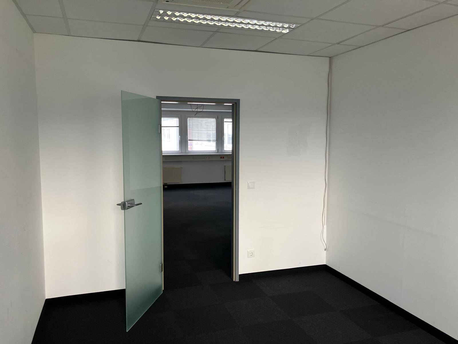 Leeres Büro mit dunklem Teppichboden und einer modernen Glastür, die zu einem weiteren Raum führt.