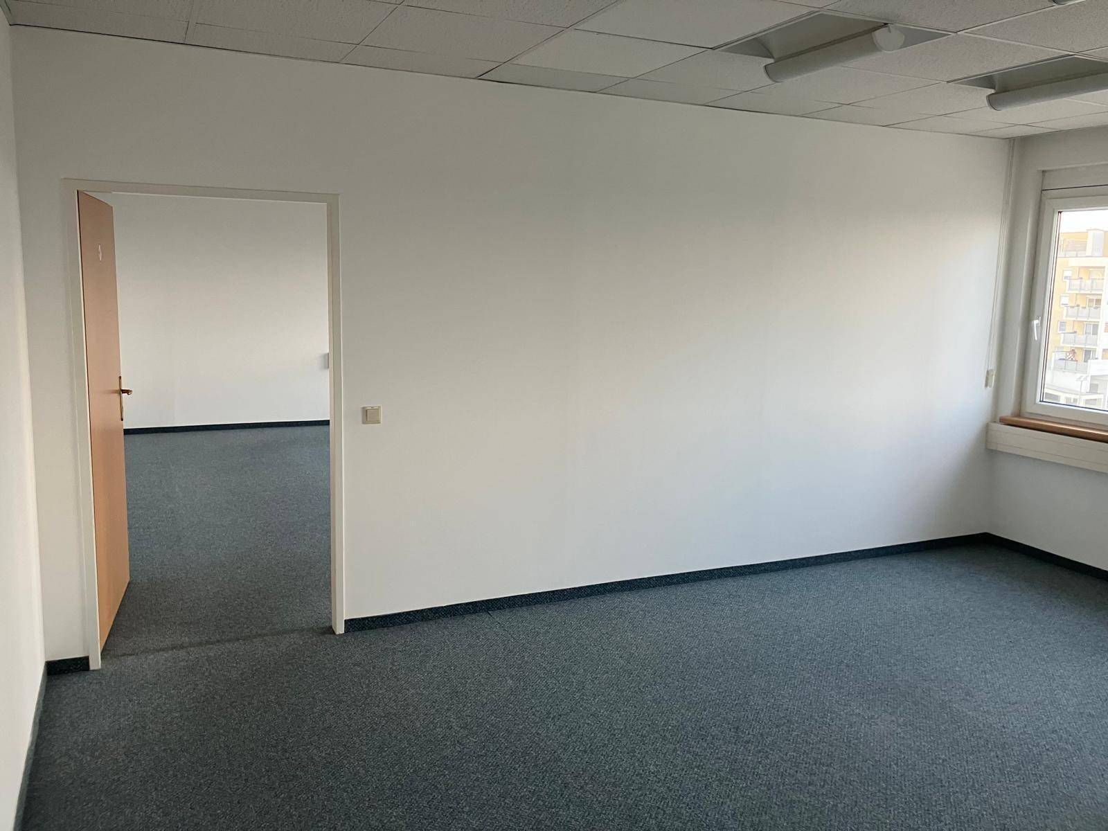 Leerer Büroraum mit Teppichboden, weißen Wänden und einem Fenster mit Stadtblick.