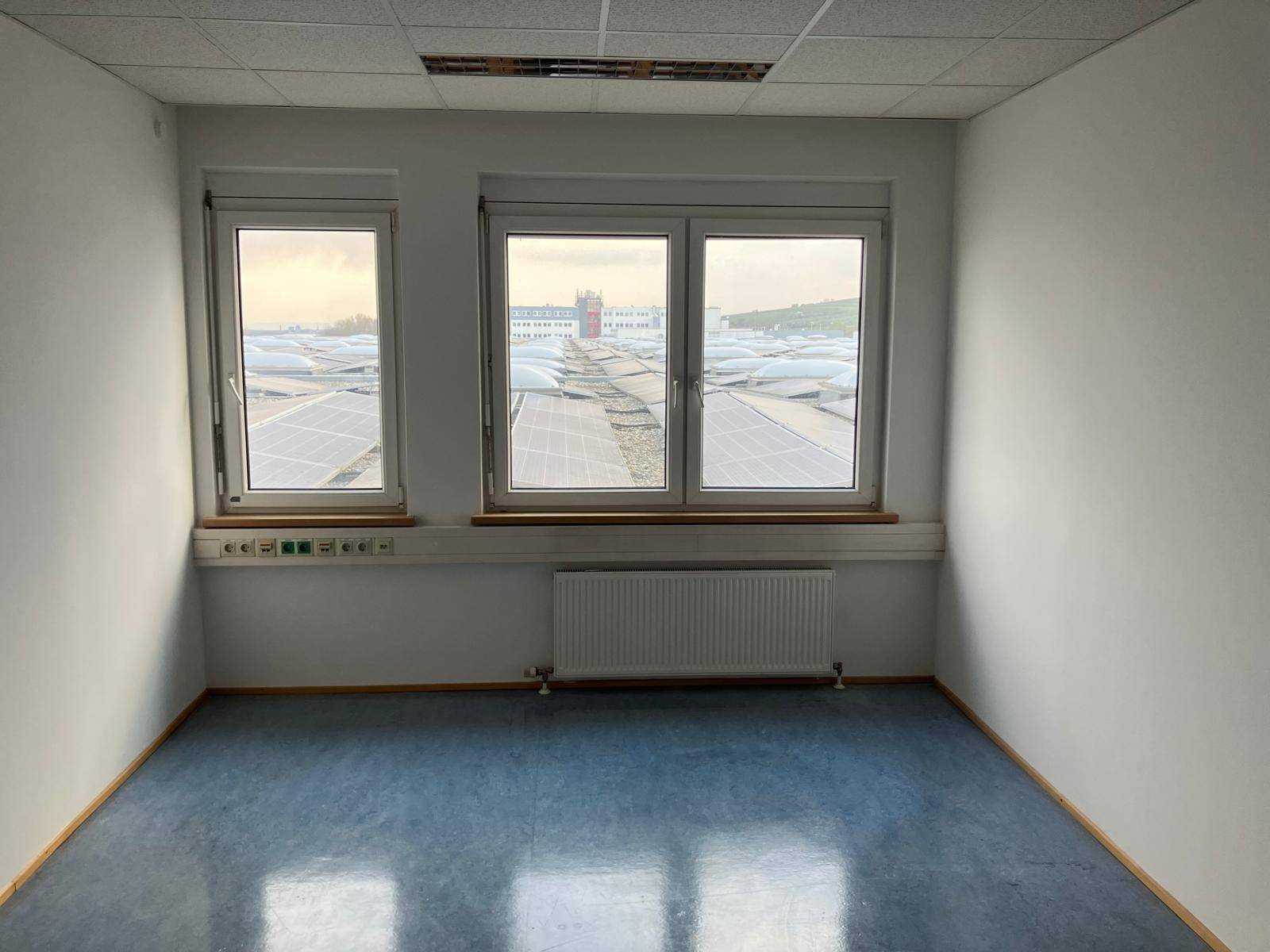 Kleines Büro mit drei Fenstern und Blick auf Dächer mit Photovoltaikanlagen.