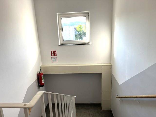 Helles Treppenhaus mit Fenster, das Tageslicht hereinlässt, und einem Feuerlöscher an der Wand.
