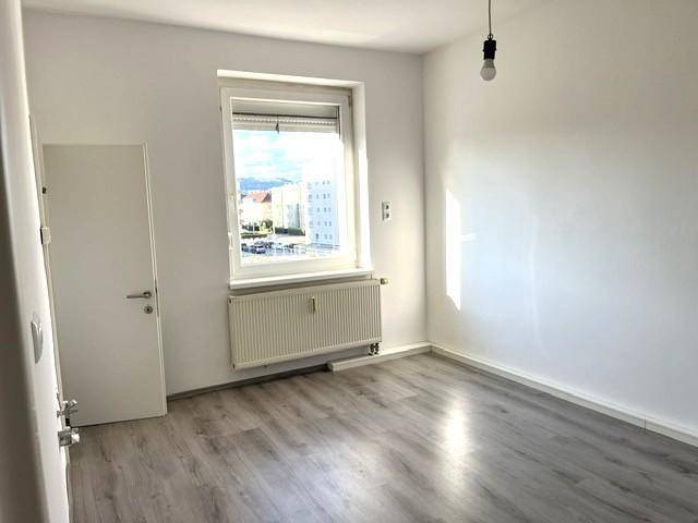 Geräumiges, helles Zimmer mit großem Fenster, Heizkörper und dunklem Laminatboden.