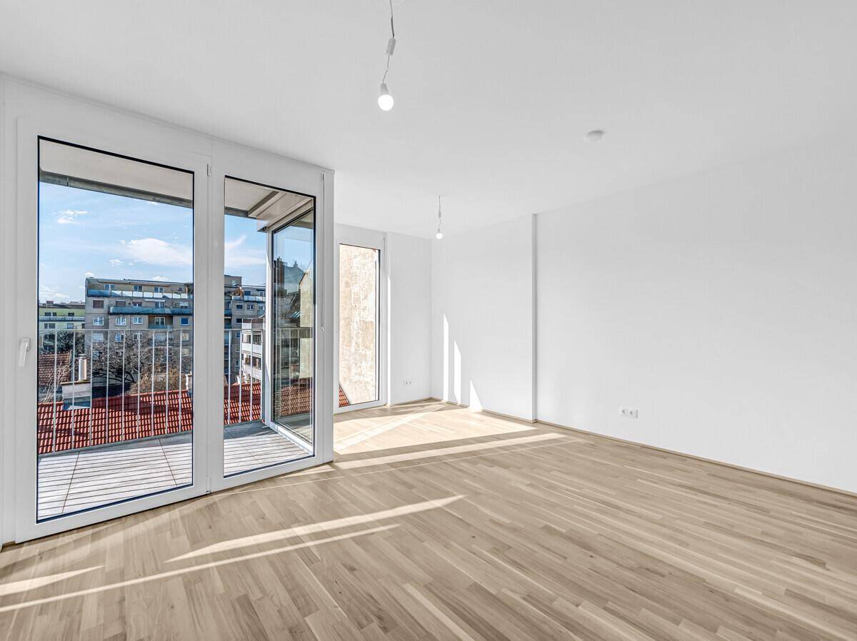 Lichtdurchfluteter, leerer Raum mit Holzboden und großen Fenstern, die Zugang zu einem Balkon bieten.