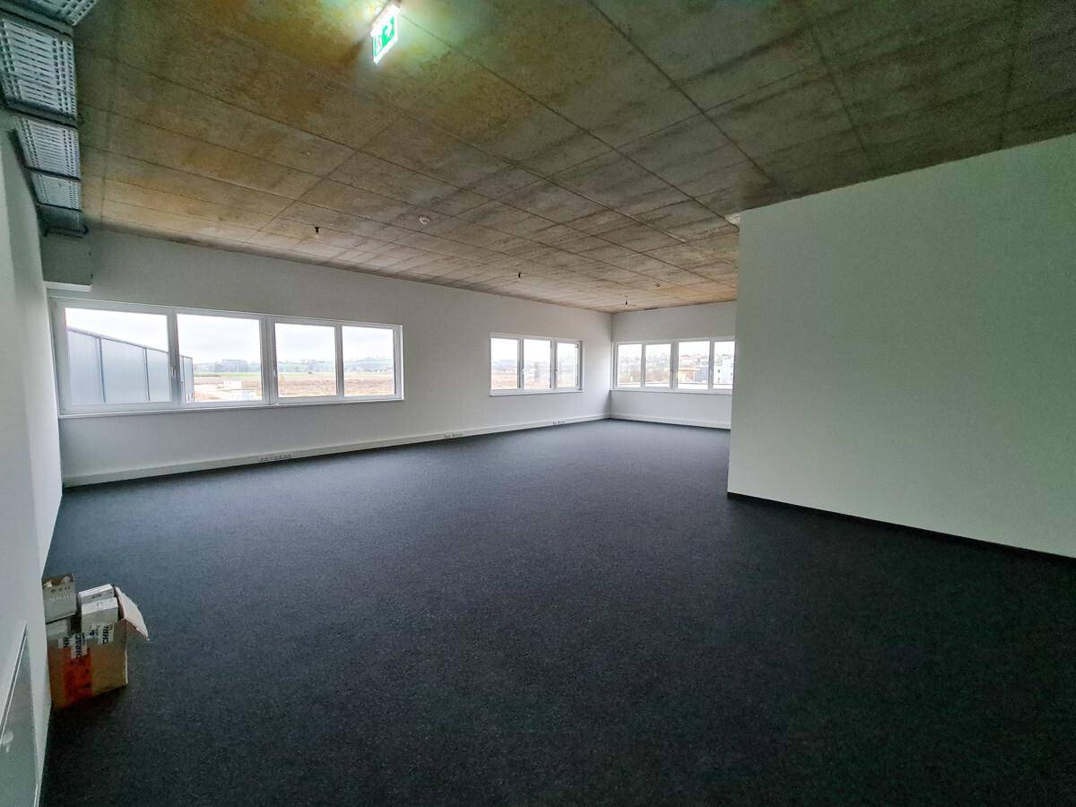 Geräumiges, leeres Büro mit dunklem Teppichboden, vielen Fenstern und einer Sichtbetondecke.