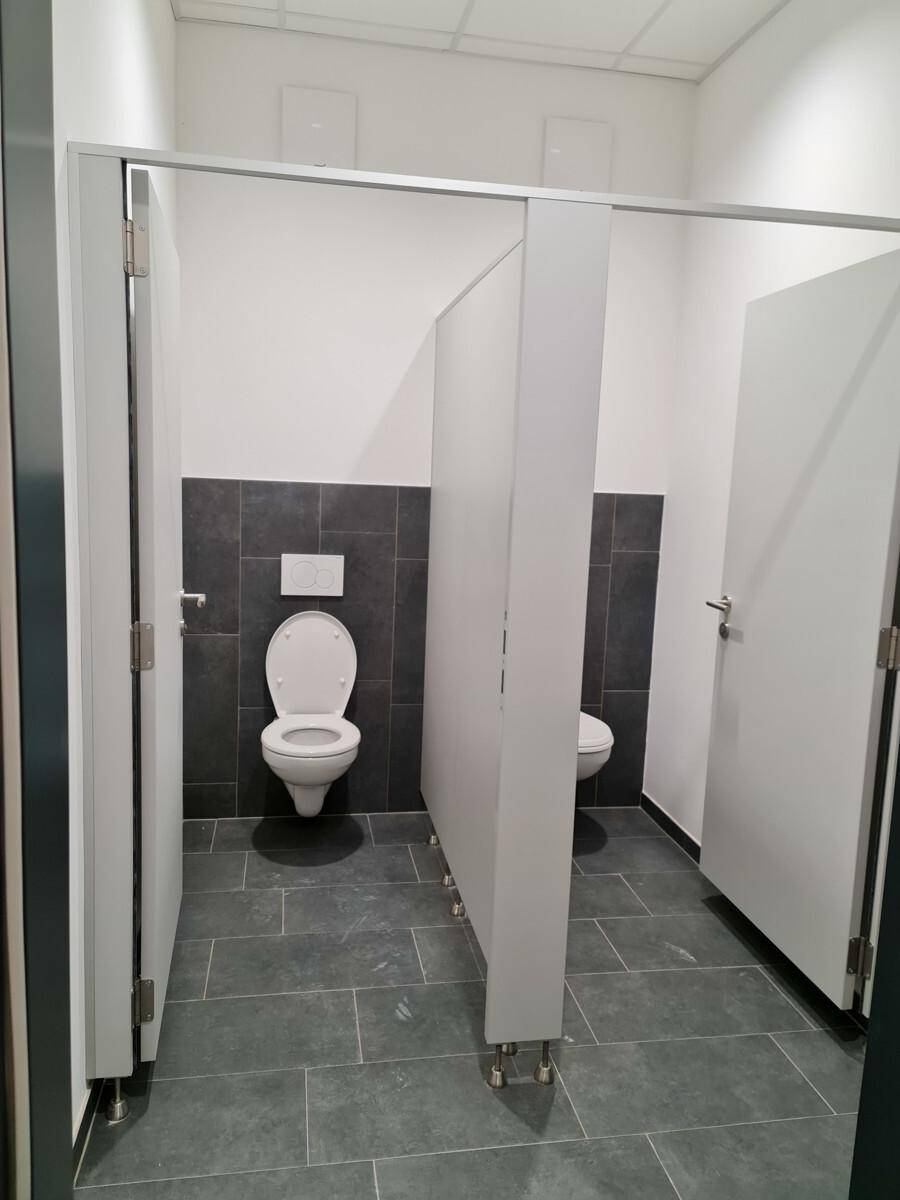 Moderner Sanitärbereich mit mehreren Kabinen, wandhängenden Toiletten und grauen Fliesen an Wänden und Boden.