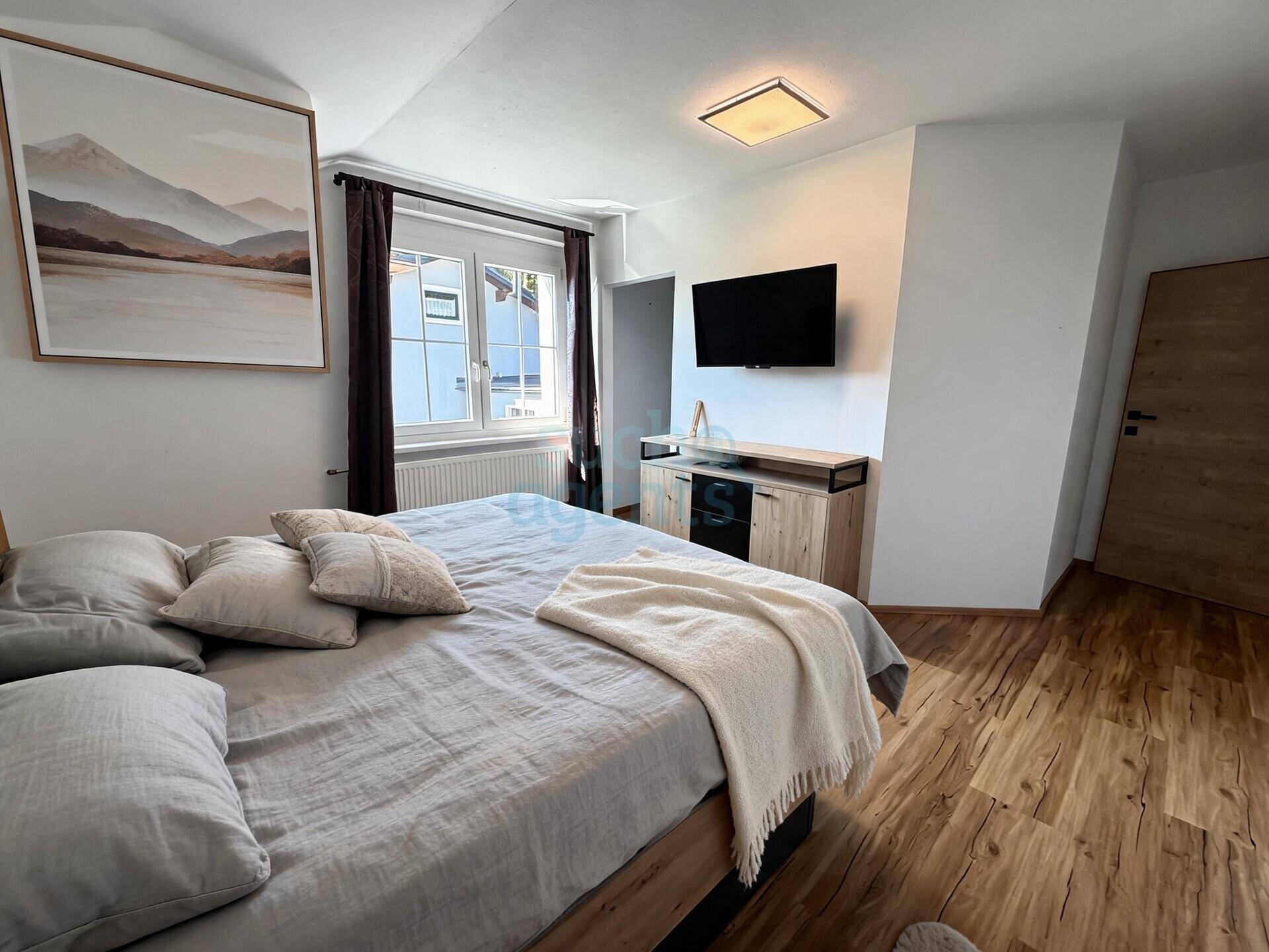 Helles Schlafzimmer mit Holzboden, großem Bett, Wandbild und Blick nach draußen durch das Fenster.