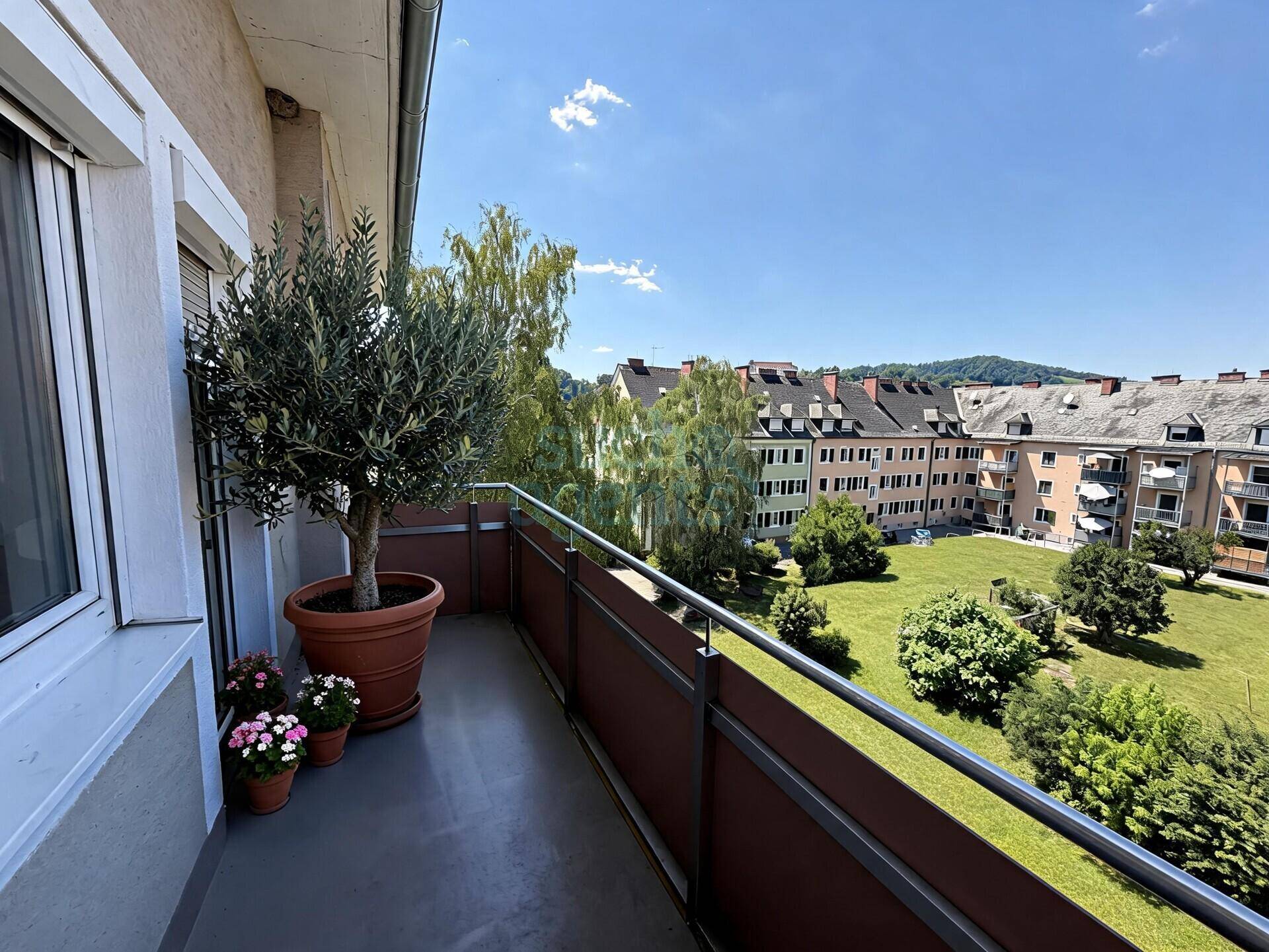 Sonniger Balkon mit Topfpflanzen und einem weiten Blick über die umliegende Wohngegend und viel Grün.
