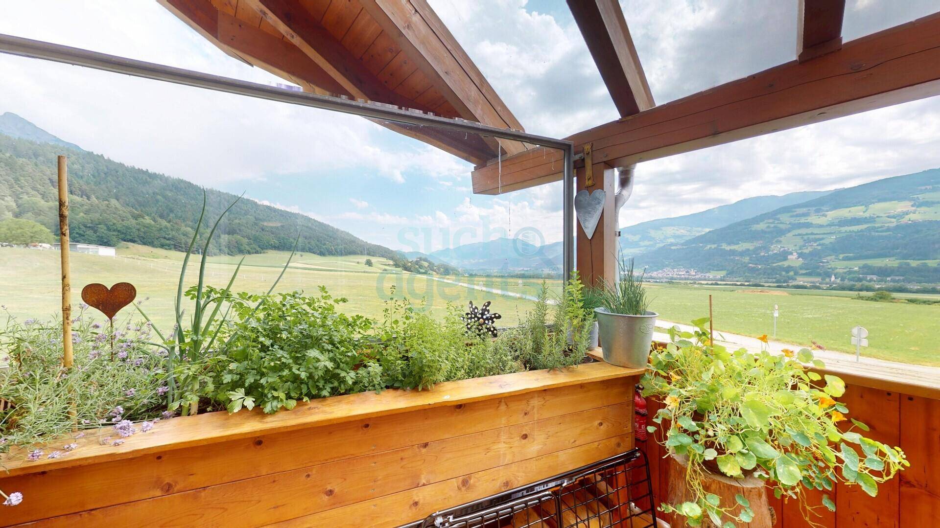 Überdachter Balkon mit Holzboden, Pflanzen und weitem Blick auf die umliegende Berglandschaft.