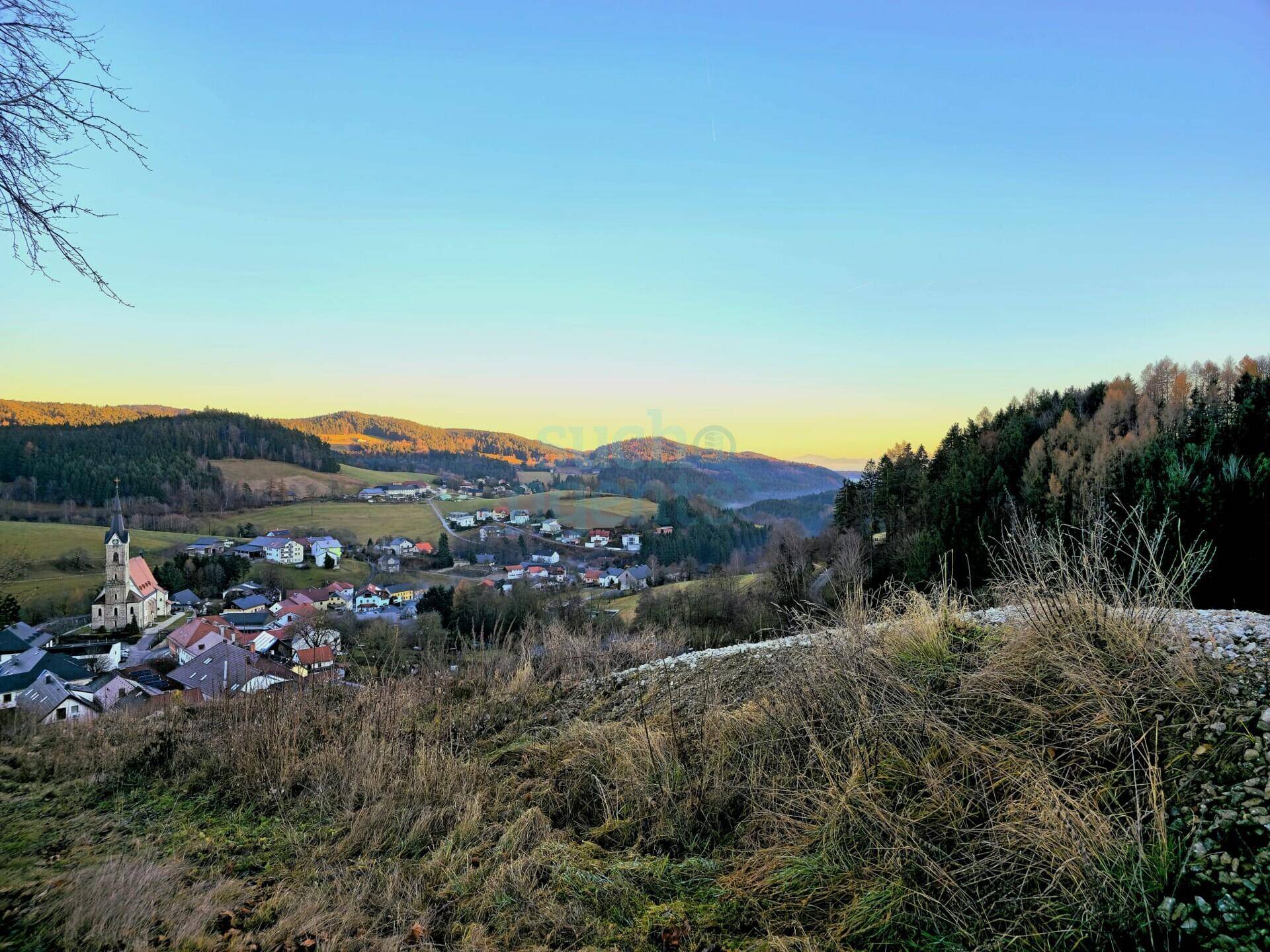 Dorf in einem Tal bei Sonnenuntergang oder Sonnenaufgang, umgeben von Hügeln und Wäldern.