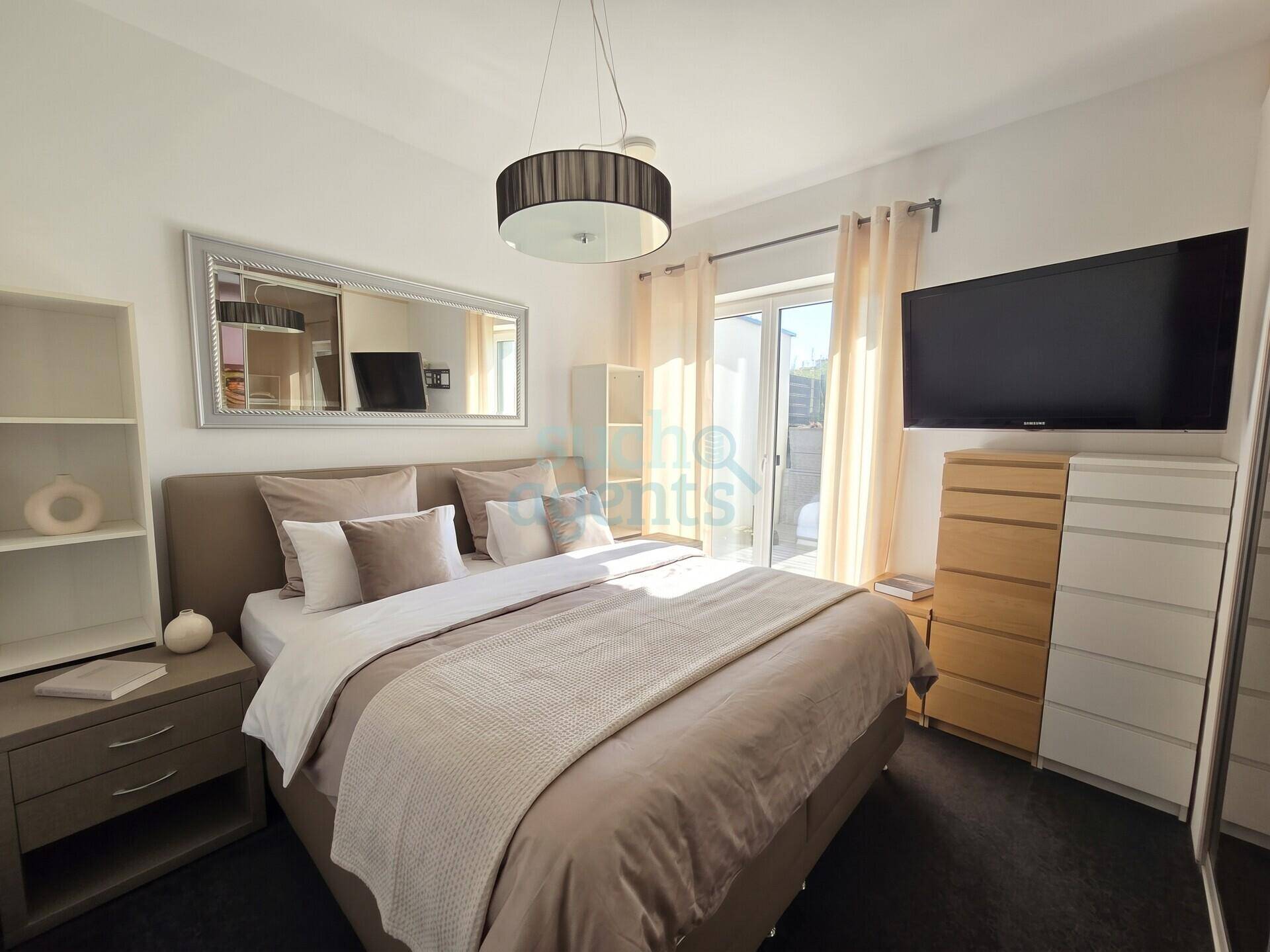 Helles Schlafzimmer mit großem Bett, Wandfernseher und direktem Zugang zu einem Balkon oder einer Terrasse.