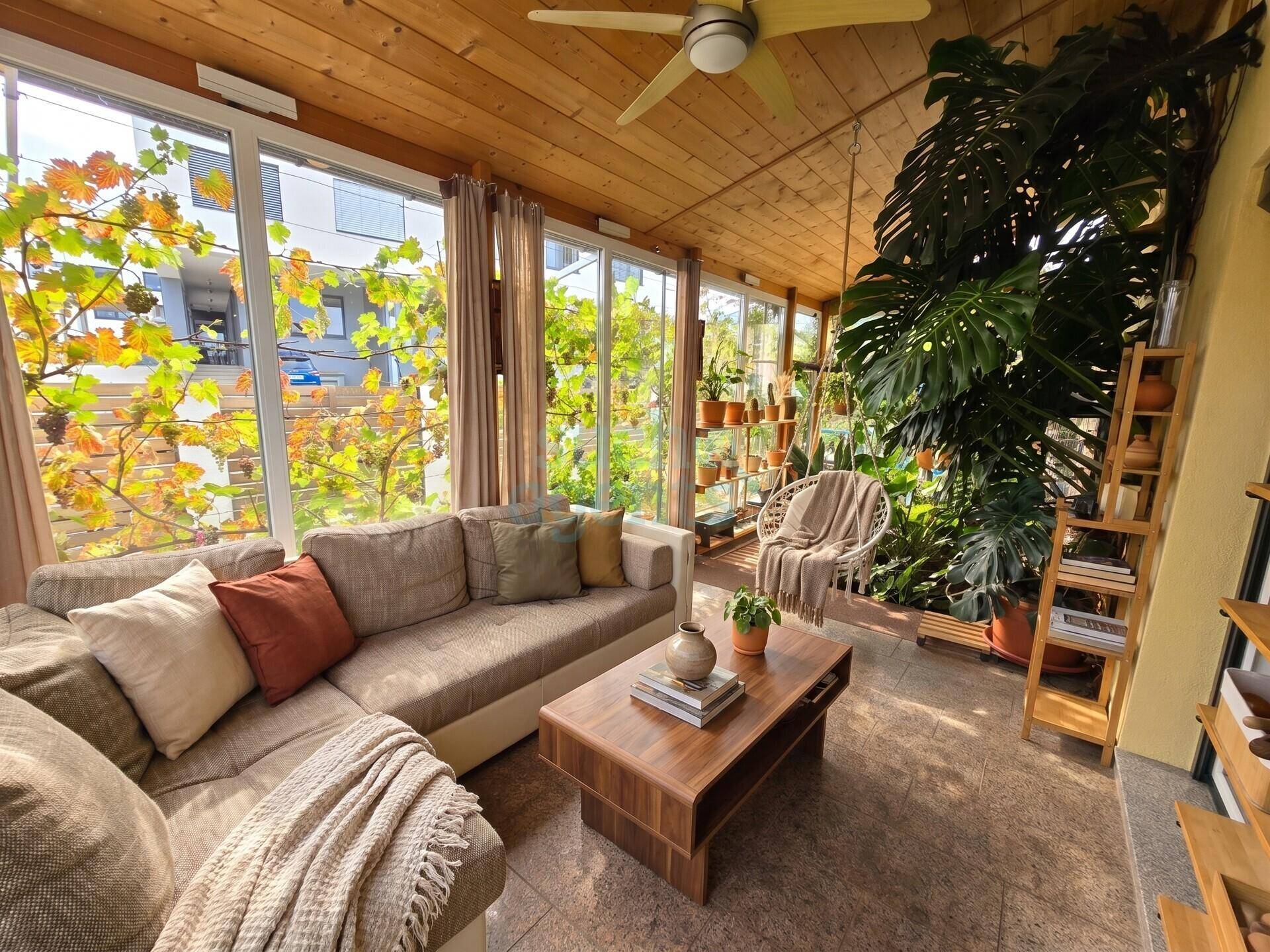 Heller Wintergarten mit großen Fenstern, bequemer Couch, Deckenventilator und vielen grünen Pflanzen.