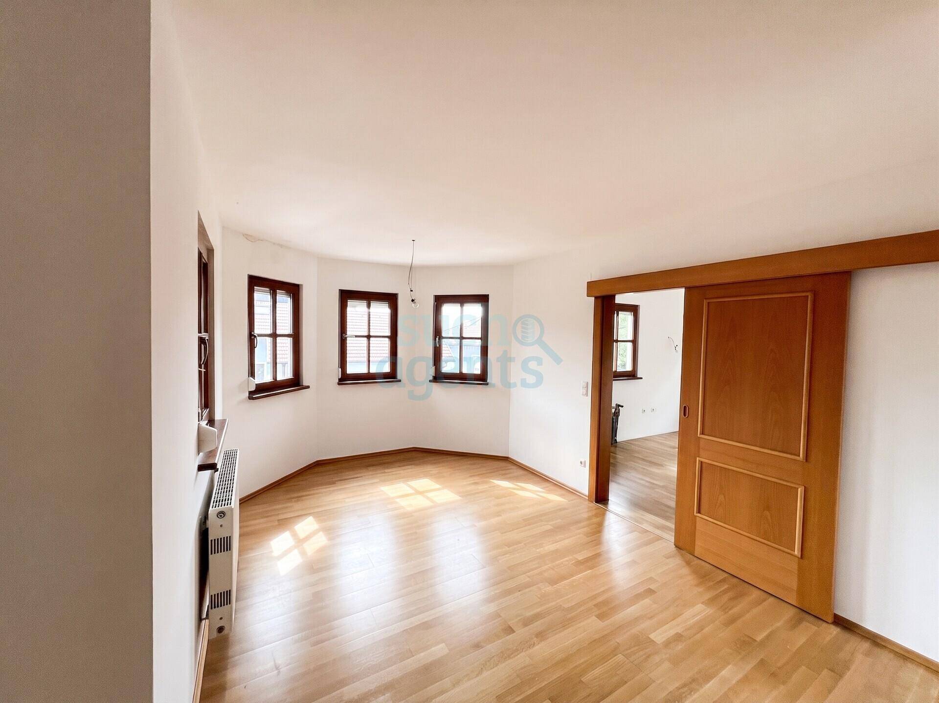 Geräumiger, heller Raum mit Holzboden, mehreren Fenstern und einer Schiebetür zu einem weiteren Zimmer.