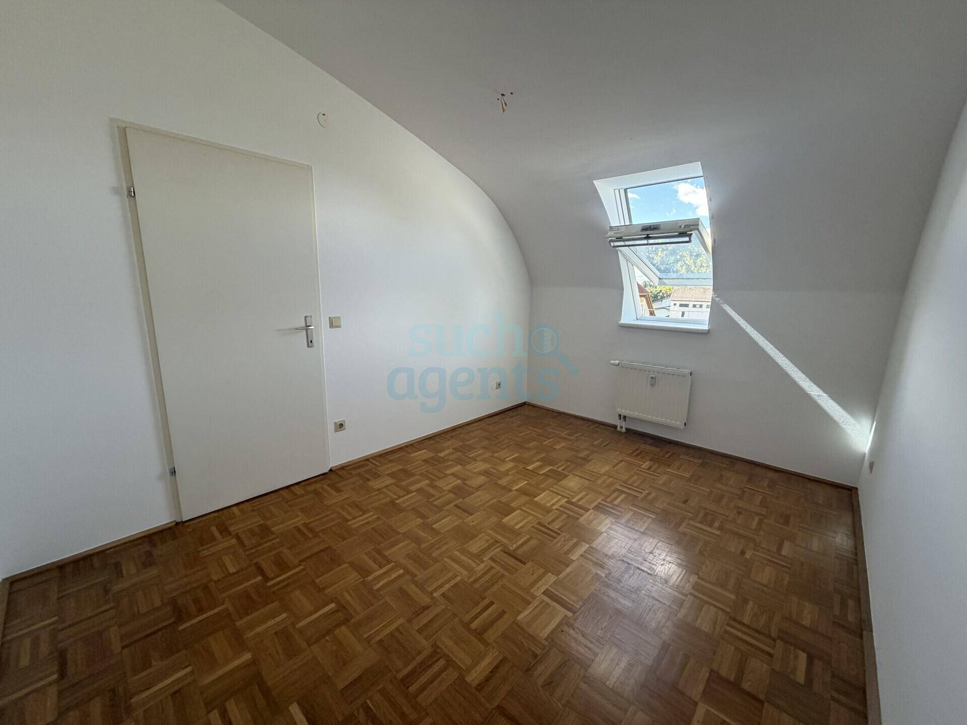 Helles Schlafzimmer im Dachgeschoss mit Parkettboden, weiß gestrichenen Wänden und einem Dachfenster.