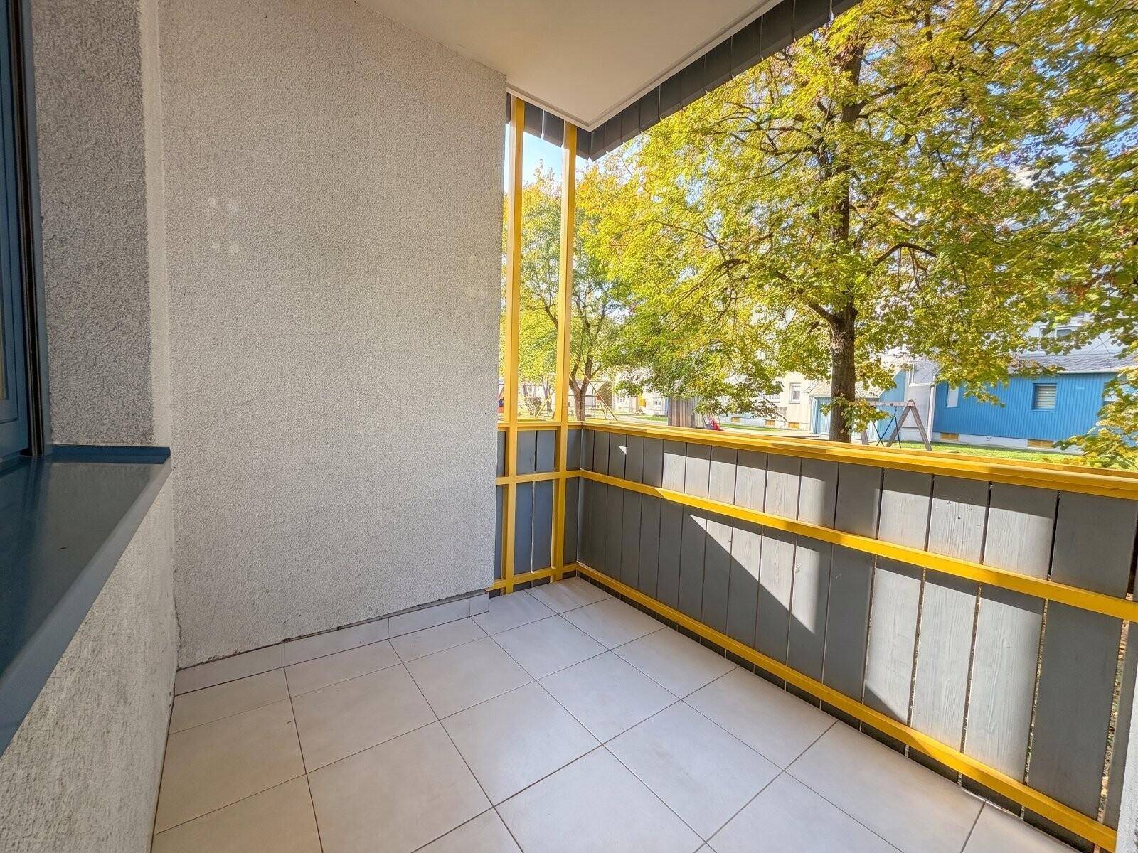 Kleiner Balkon mit Fliesenboden und Blick auf Bäume und einen Spielplatz in der Umgebung.