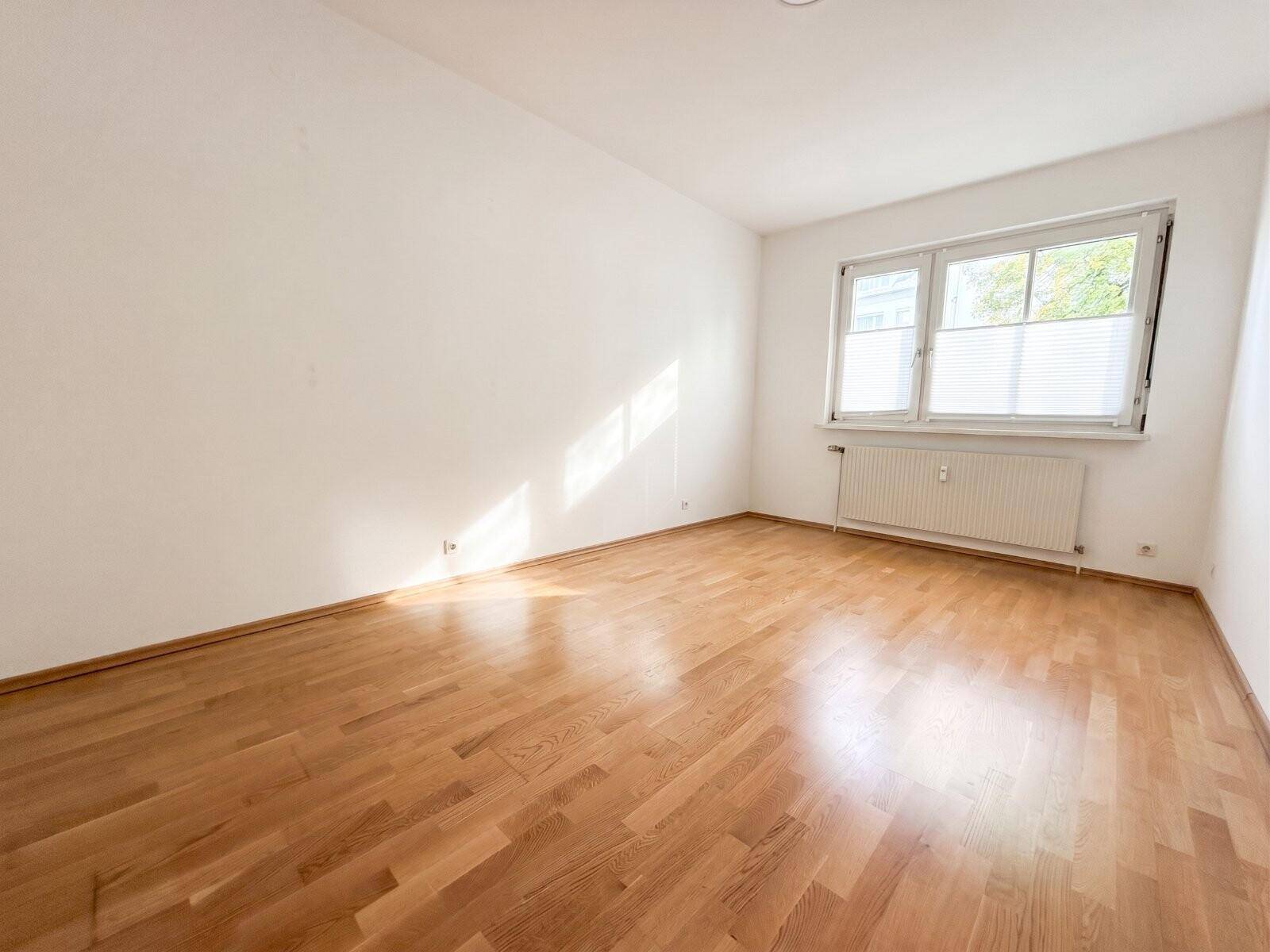 Leeres Zimmer mit Parkettboden und großem Fenster, ideal als Schlafzimmer oder Arbeitsbereich nutzbar.