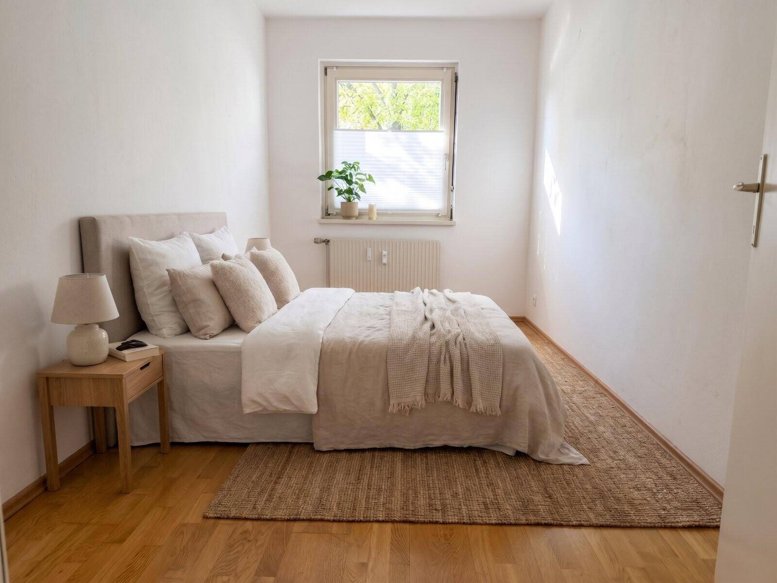 Gemütliches Schlafzimmer mit Holzboden, beigem Bett und einem Fenster, das natürliches Licht hereinlässt.