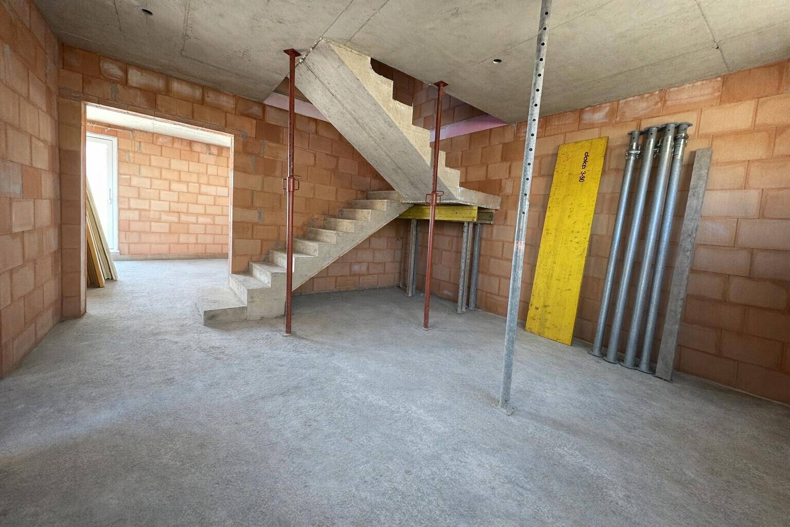 Innenansicht eines Rohbaus mit Betontreppe, Ziegelwänden und Baustützen im Eingangsbereich.