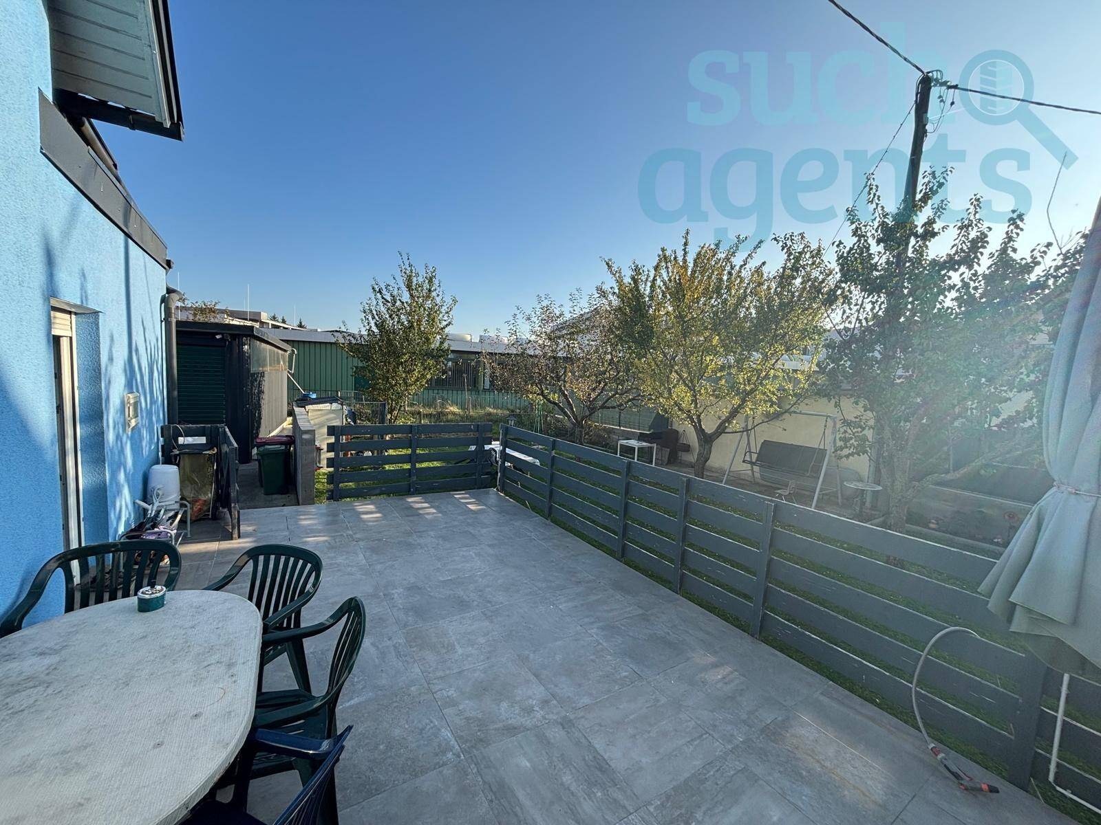 Sonniger Außenbereich mit gepflasterter Terrasse, Gartenzaun und Sitzgelegenheiten.