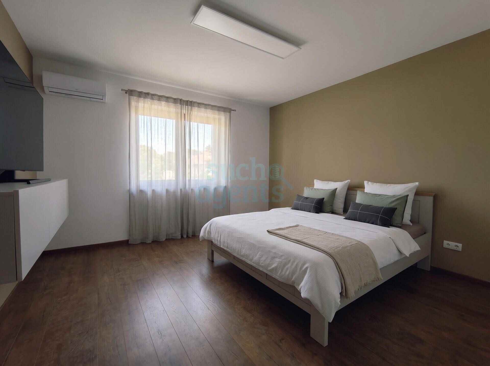 Helles Schlafzimmer mit Holzboden, großem Fenster und Klimaanlage für angenehme Temperaturen.