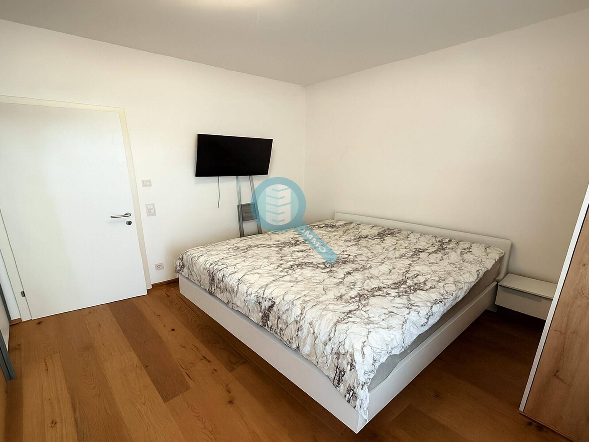 Helles Schlafzimmer mit Holzboden, Doppelbett und einem an der Wand montierten Fernseher.