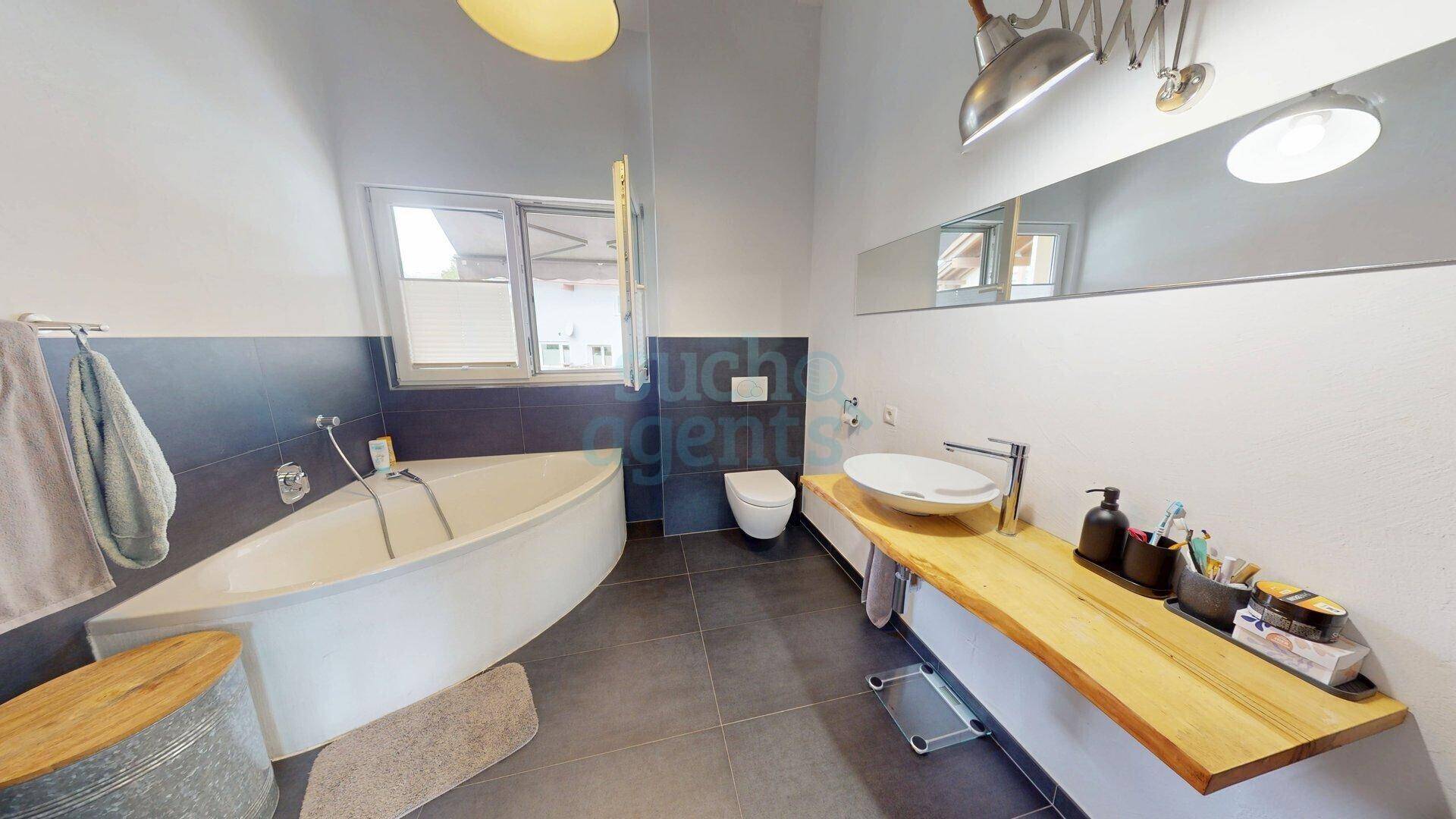Modernes Badezimmer mit Eckbadewanne, Holzwaschtisch, Wand-WC und Fenster für Tageslicht.