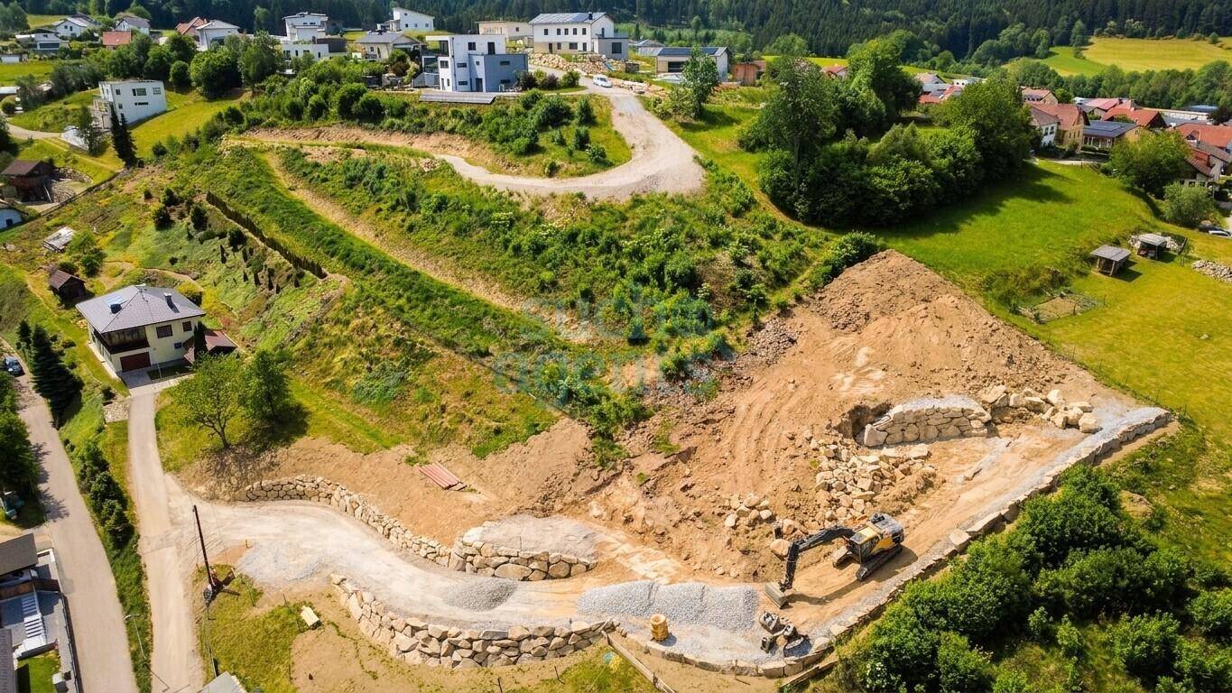 Luftaufnahme einer Baustelle auf einem Hügel mit Erdarbeiten, Stützmauern und einem Bagger.